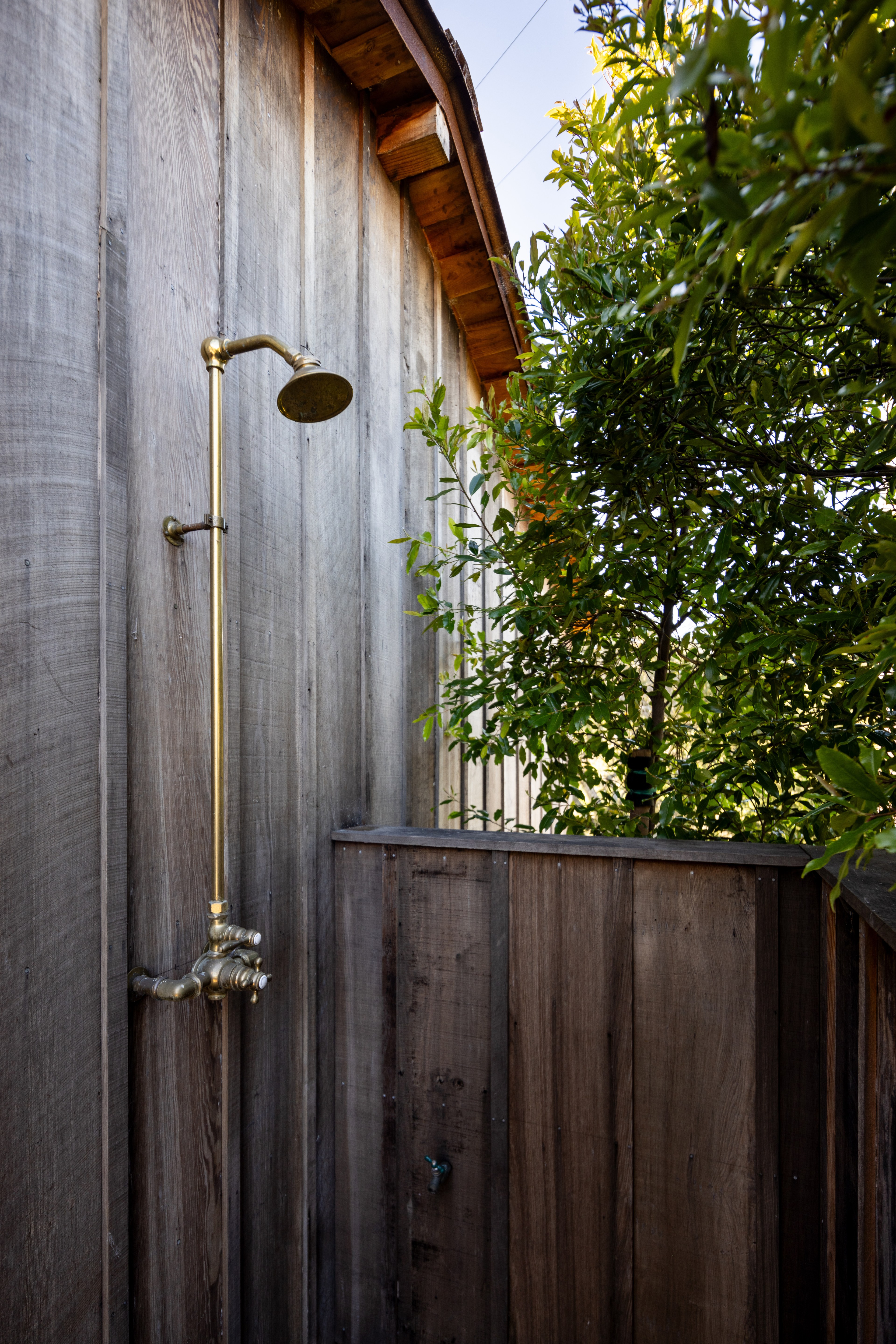 A rustic outdoor shower is set against a weathered wooden wall with brass fixtures. It's partially enclosed by more wood and surrounded by green foliage.