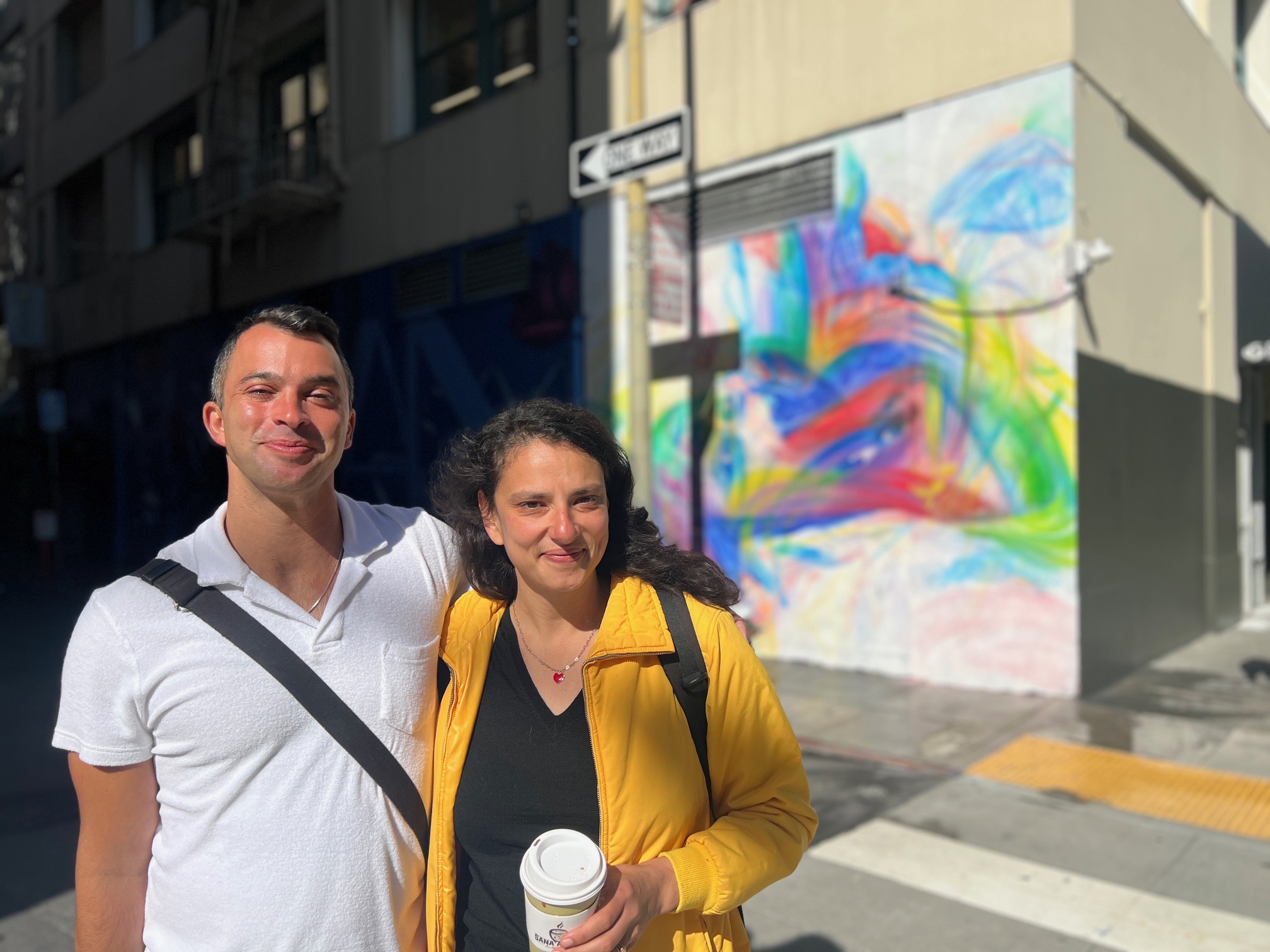 A smiling man and woman on a sunny street; colorful mural in the background.