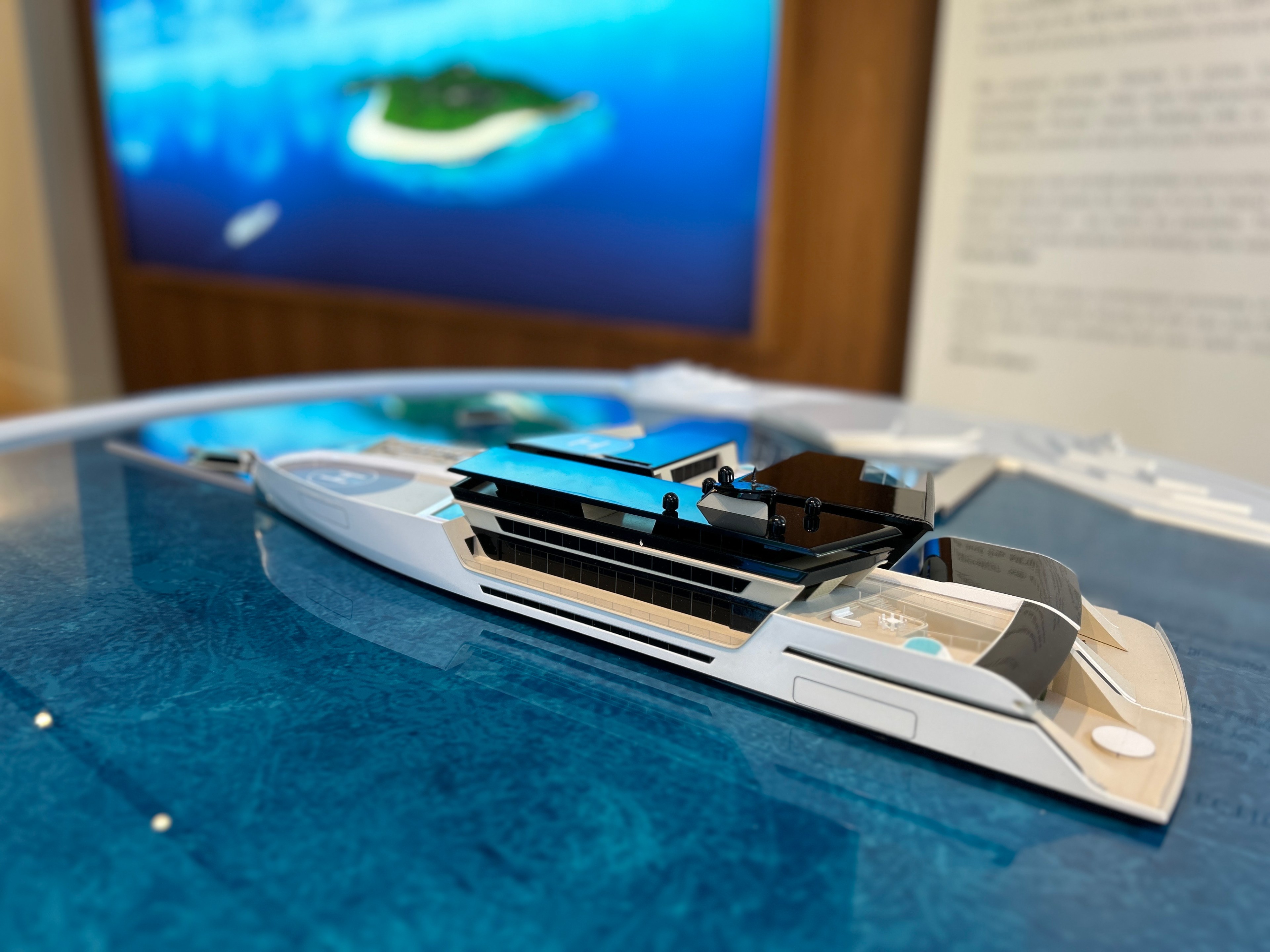 a model of a superyacht on a reflective table meant to evoke the ocean, with a screen showing a tropical island in the background