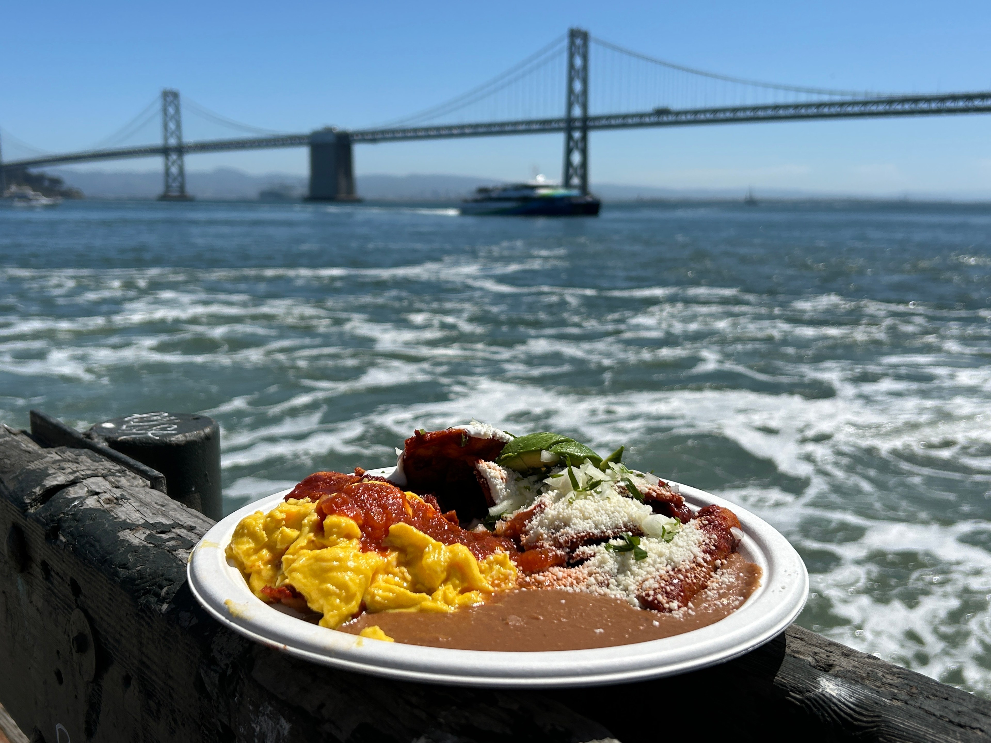 a plate of chilaquilles with eggs and refried beans sits on a rail at the waterfront with choppy SF Bay and the Bay Bridge visible