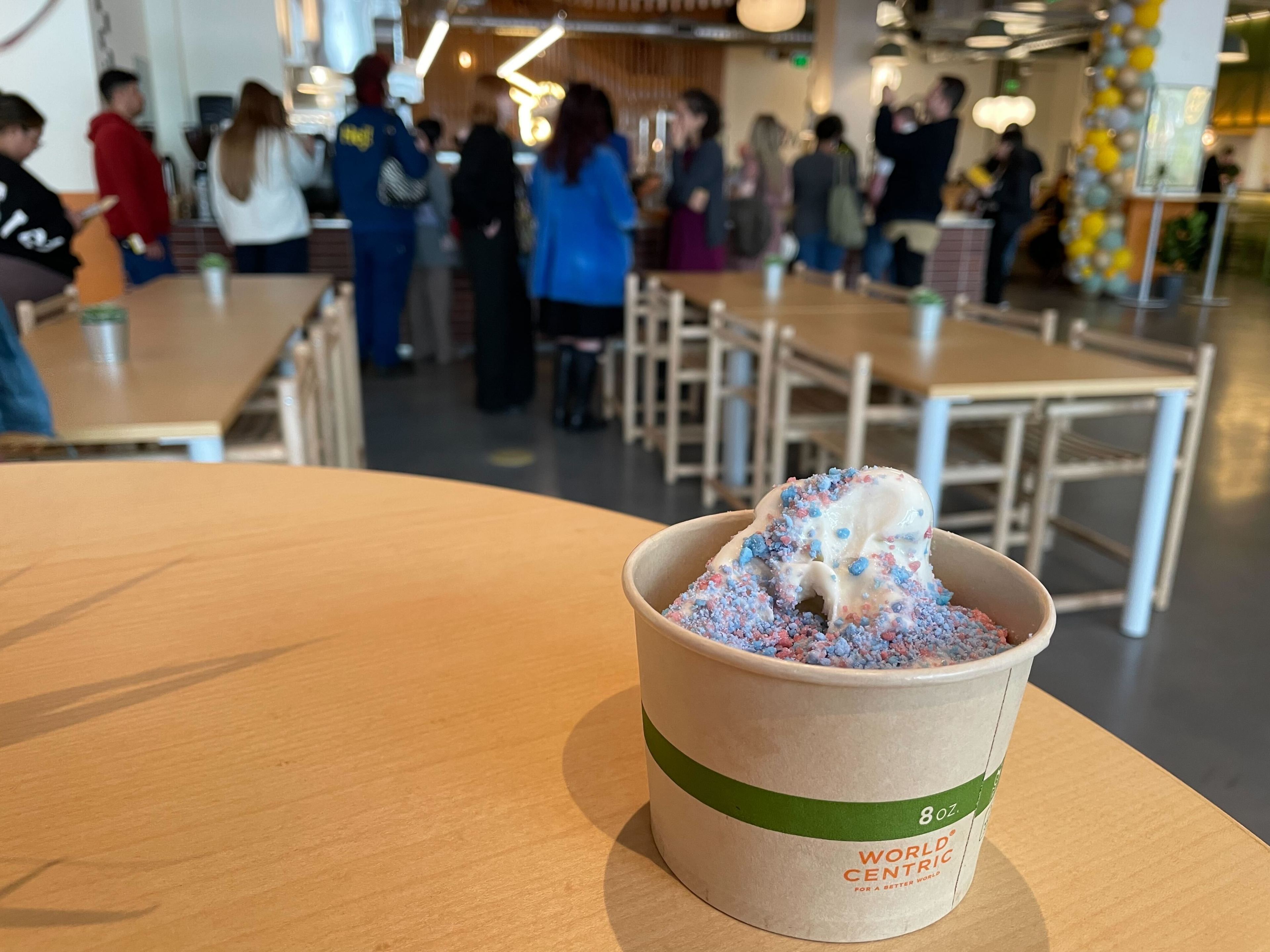 A paper cup of ice cream on a table, with sprinkles on top. Blurry background with people standing in a cafe.