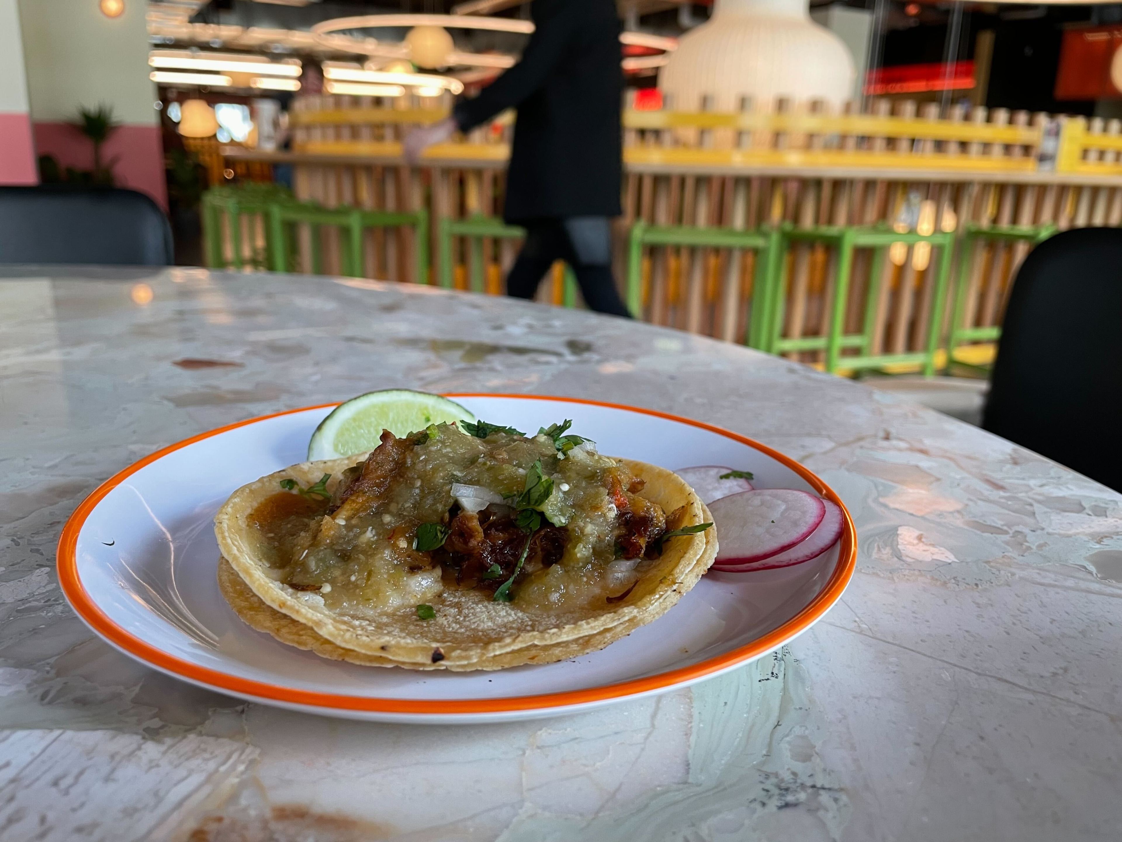 A taco with cilantro on a plate with lime, on a marble table, restaurant setting blurred in the background.