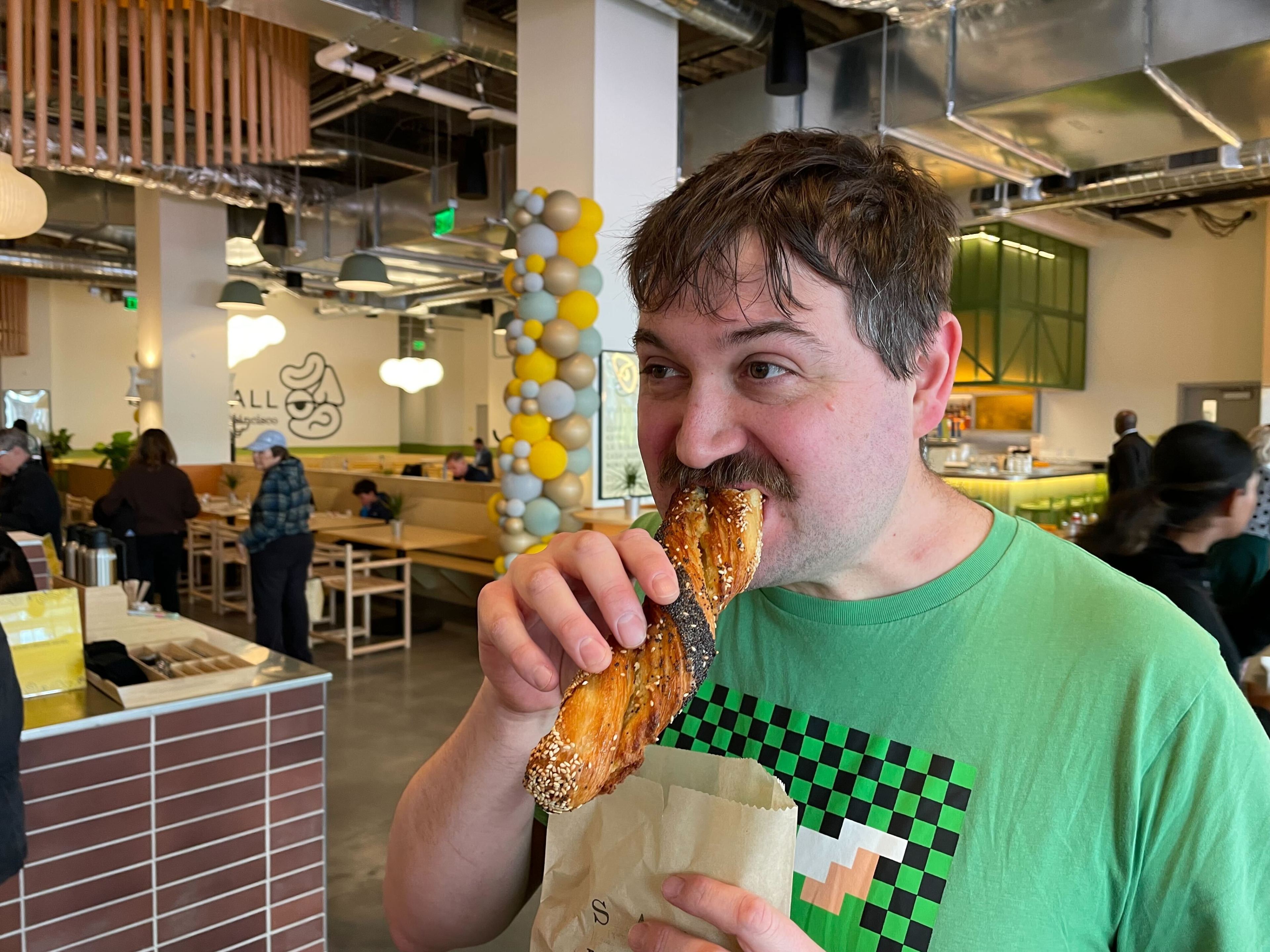 A man is biting a pretzel in a busy, modern cafe, looking pleased.