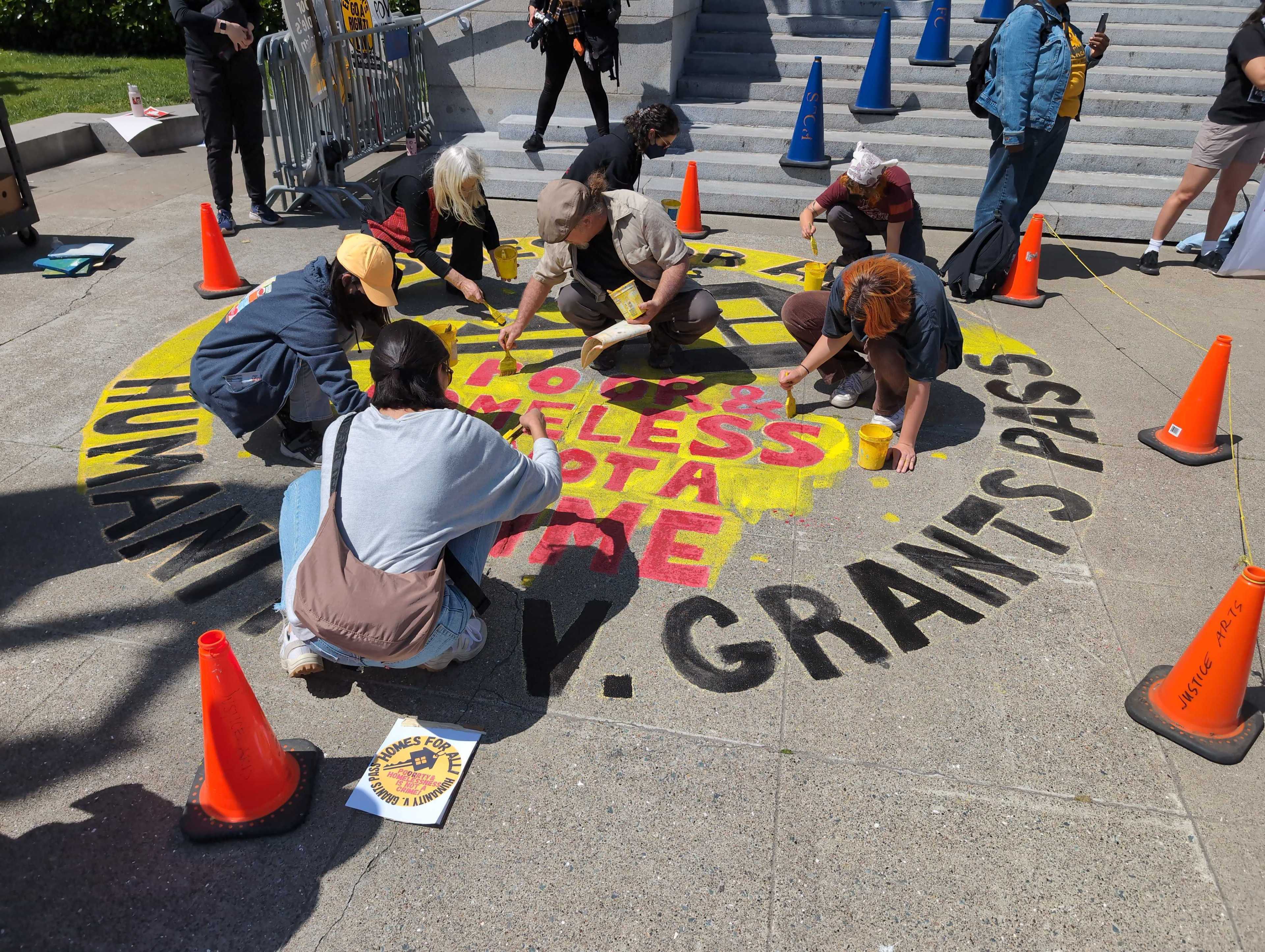 People are painting a circular street mural with the words &quot;HUMANITY NOT VANITY, SAN FRANCISCO SAYS PAST ACTS MATTER&quot; surrounded by traffic cones.