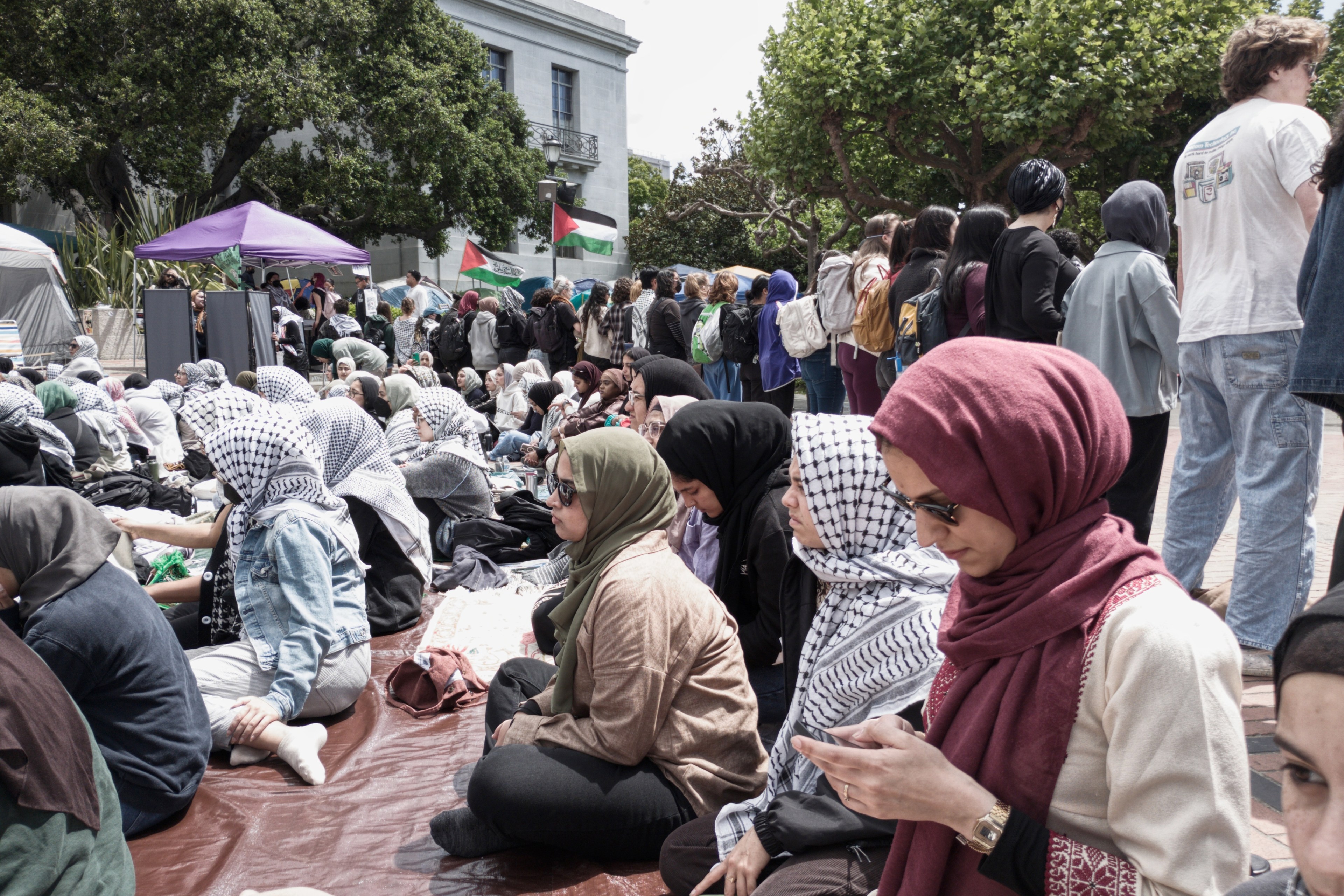 A diverse crowd is gathered outside, some sitting on the ground, others standing, many wearing headscarves, near tents and flags.