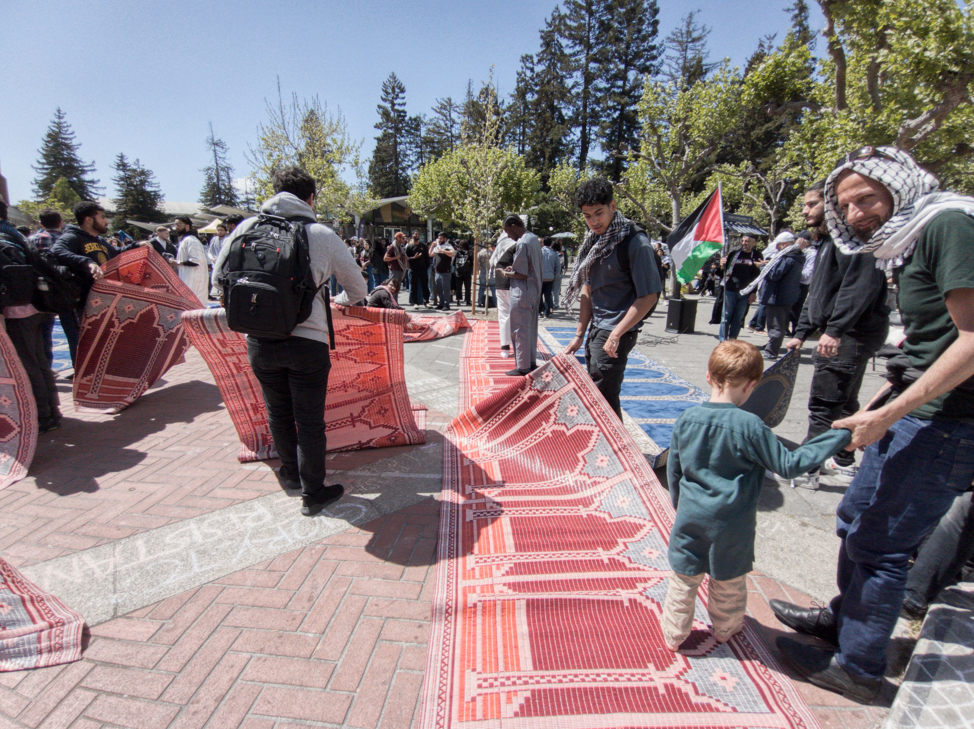 A child interacts with adults holding up a red patterned barrier at an outdoor gathering beneath trees.