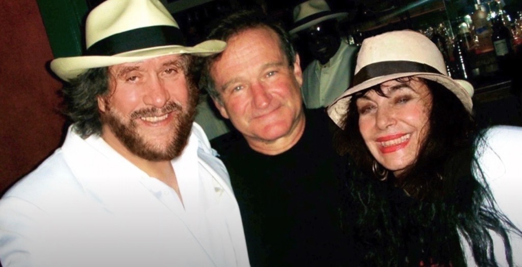 Three smiling people pose for a photo, the men wearing hats and the woman with long, dark hair. They're in front of a bar area.