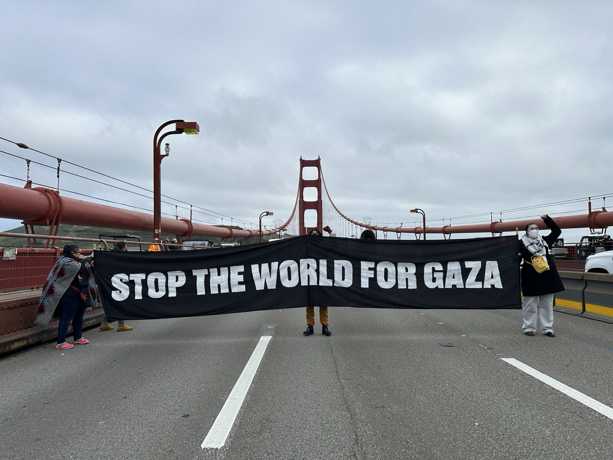 People hold a banner saying &quot;STOP THE WORLD FOR GAZA&quot; on a bridge with cloudy skies overhead.