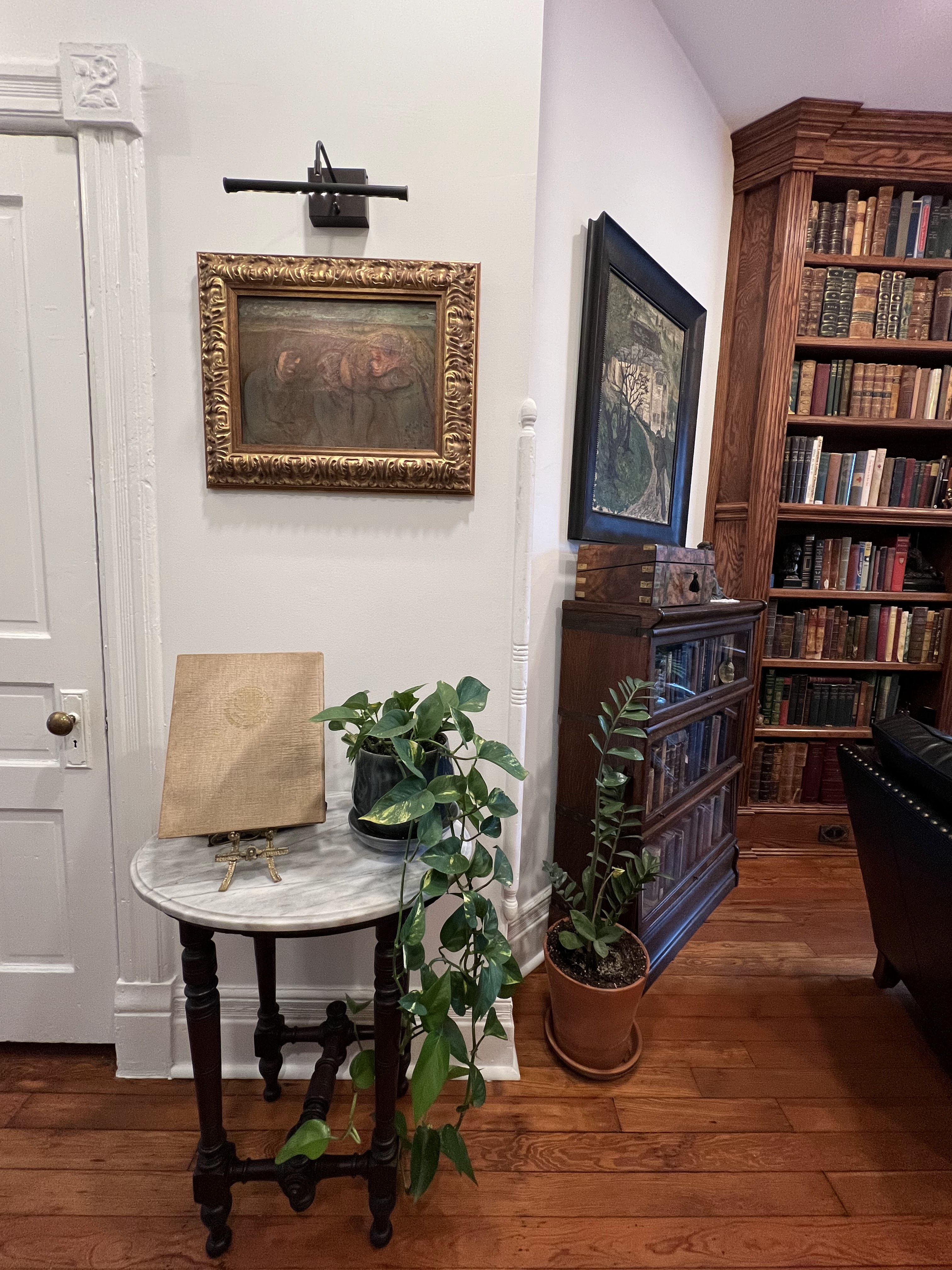 A cozy corner with a round table, plants, paintings, and a bookcase filled with books.