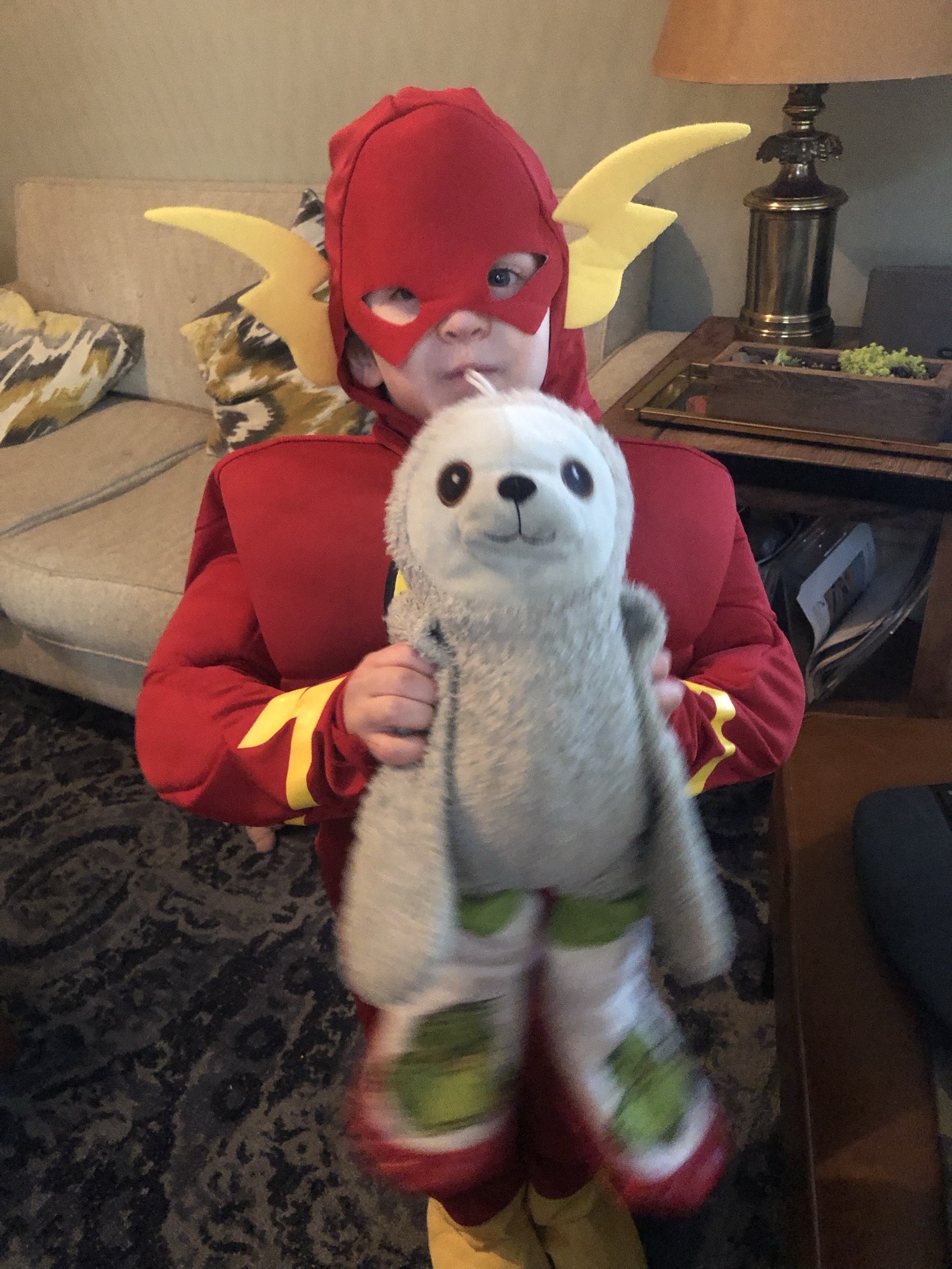 A child in a red superhero costume with a mask and flash-like emblems holds a stuffed sloth toy.