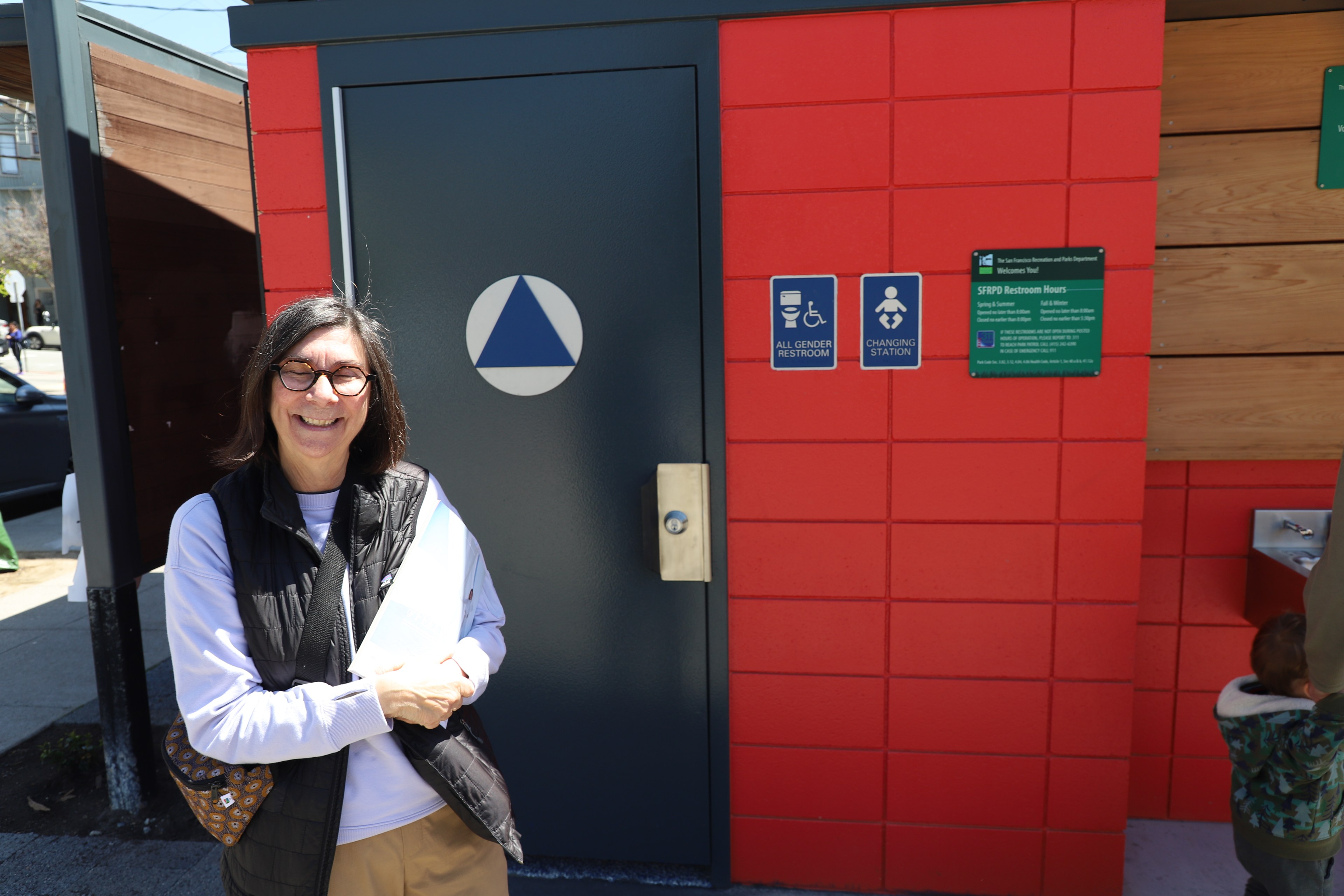 A woman stands in front of a public restroom on a sunny day.