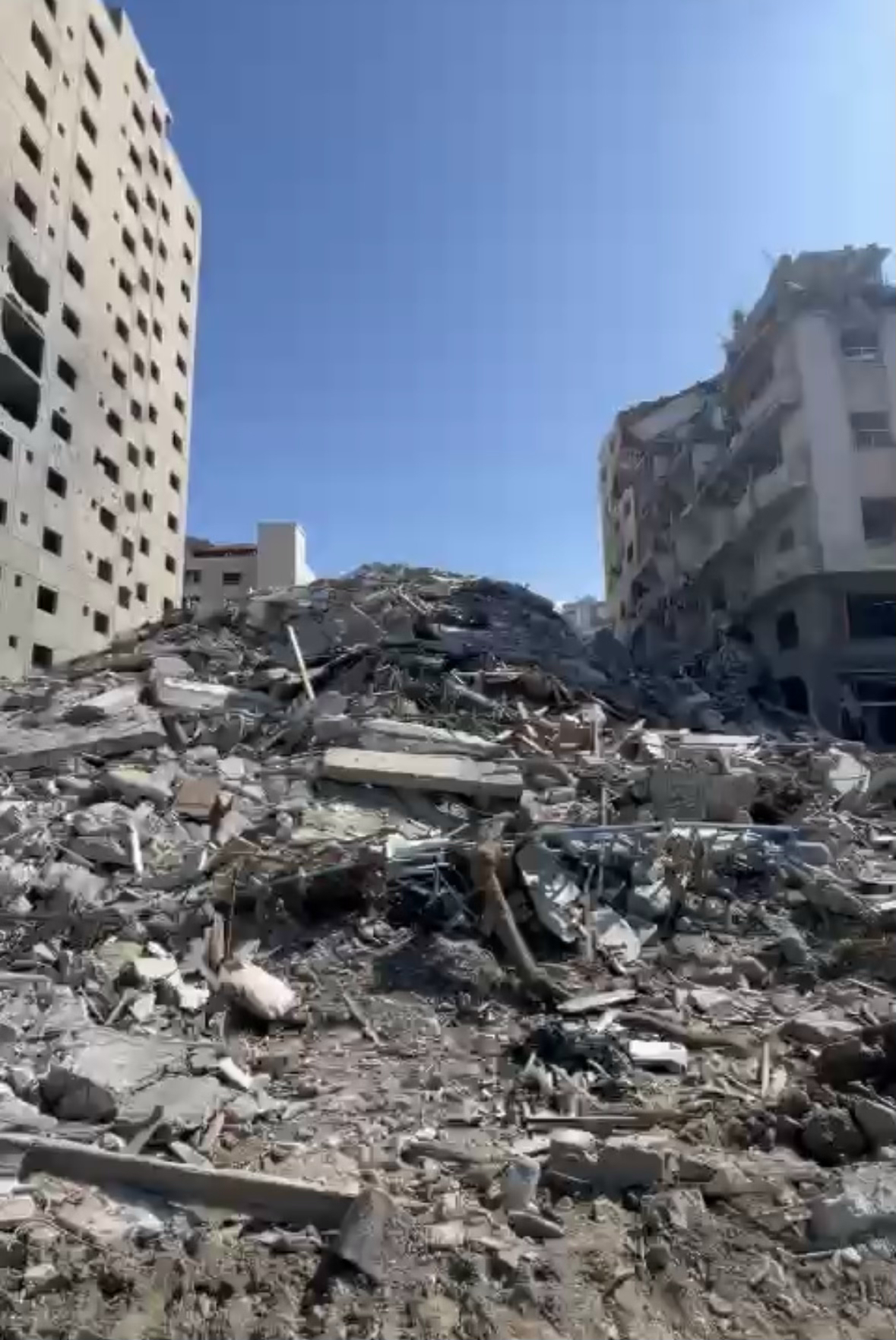 A pile of rubble with damaged buildings in the background under a clear blue sky.