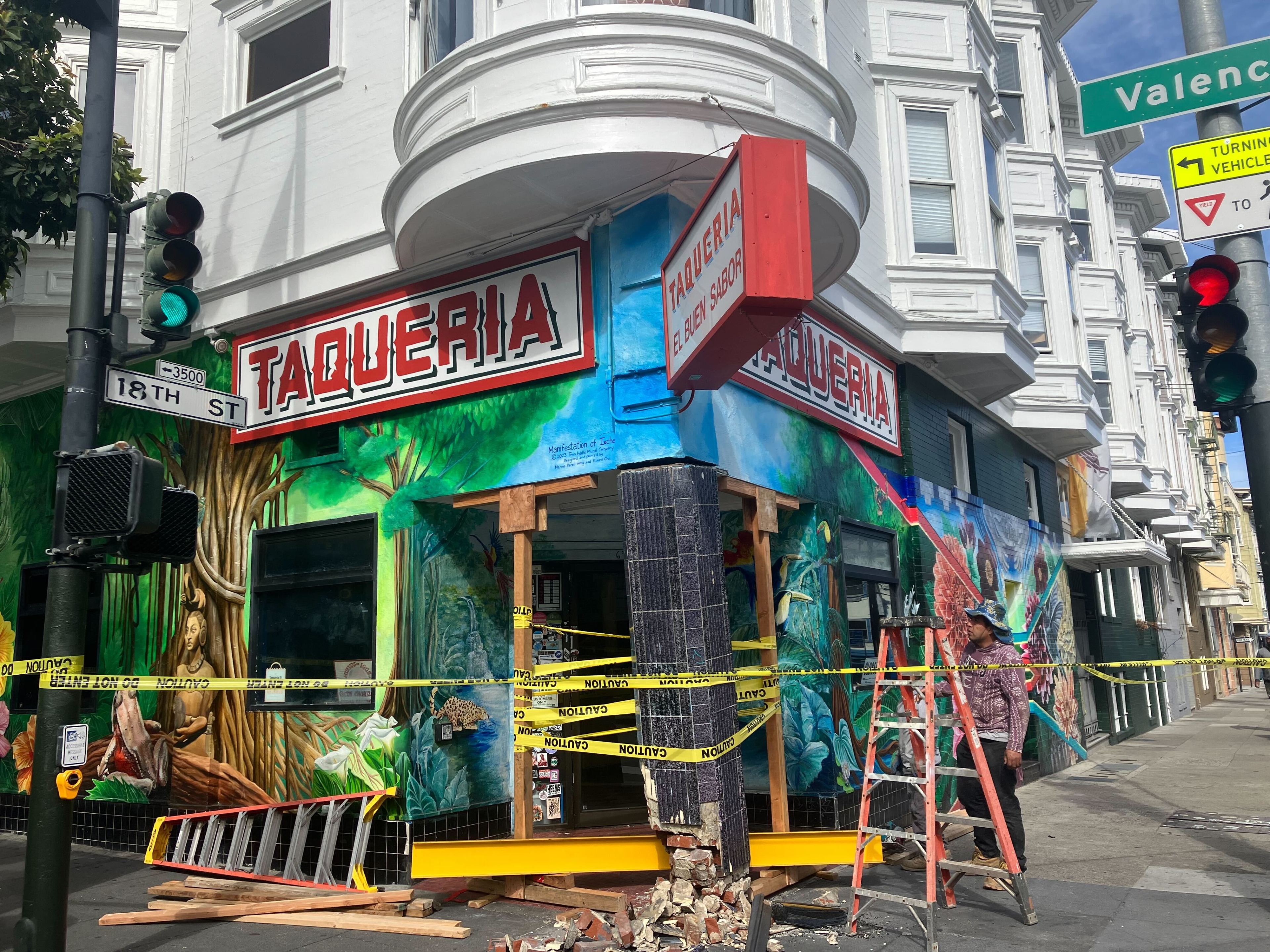 A damaged taqueria at a street corner, with colorful murals and caution tape, under repair by a person on a ladder.