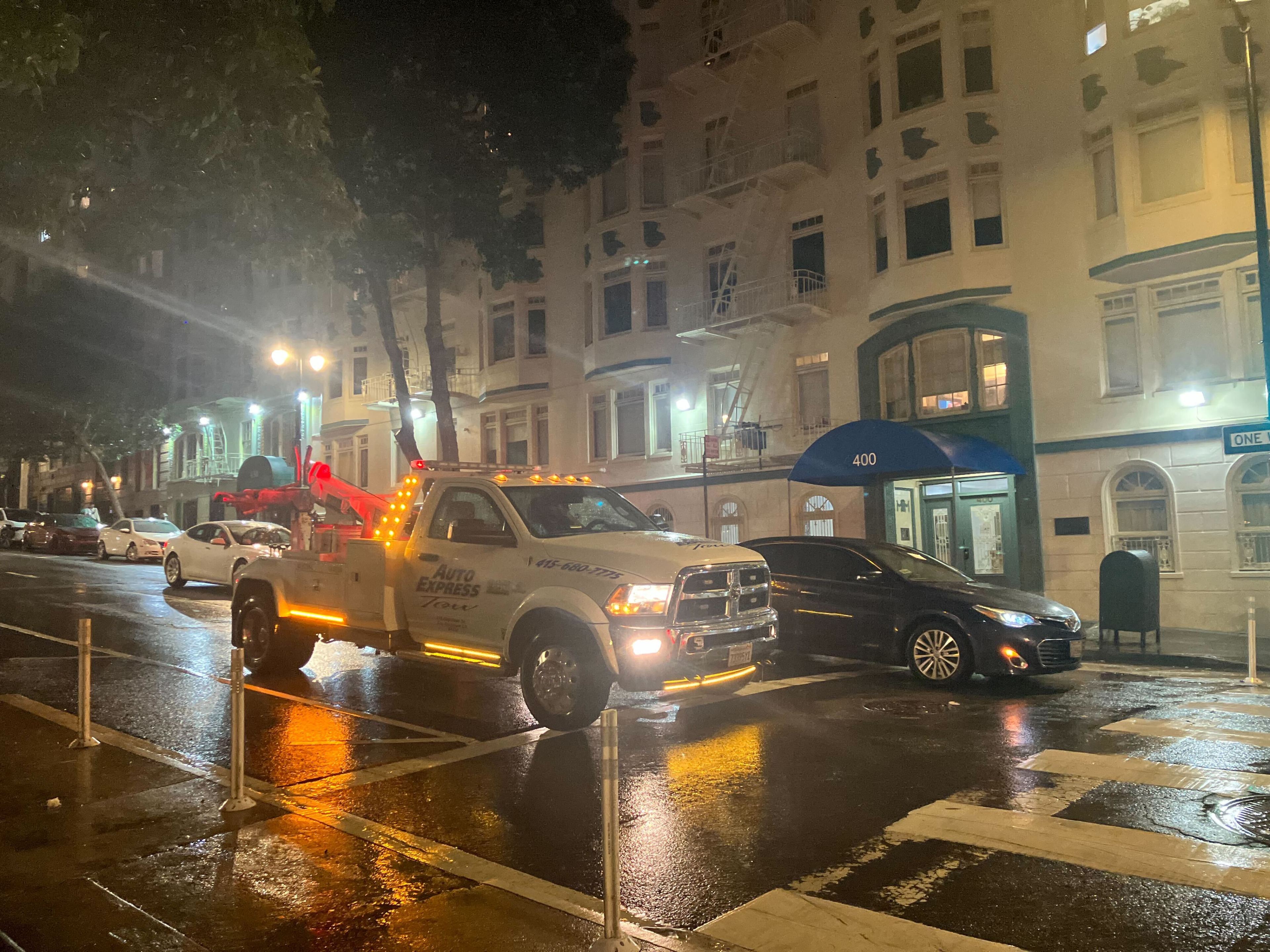 A tow truck with flashing lights is parked on a wet street at night, in front of apartment buildings.
