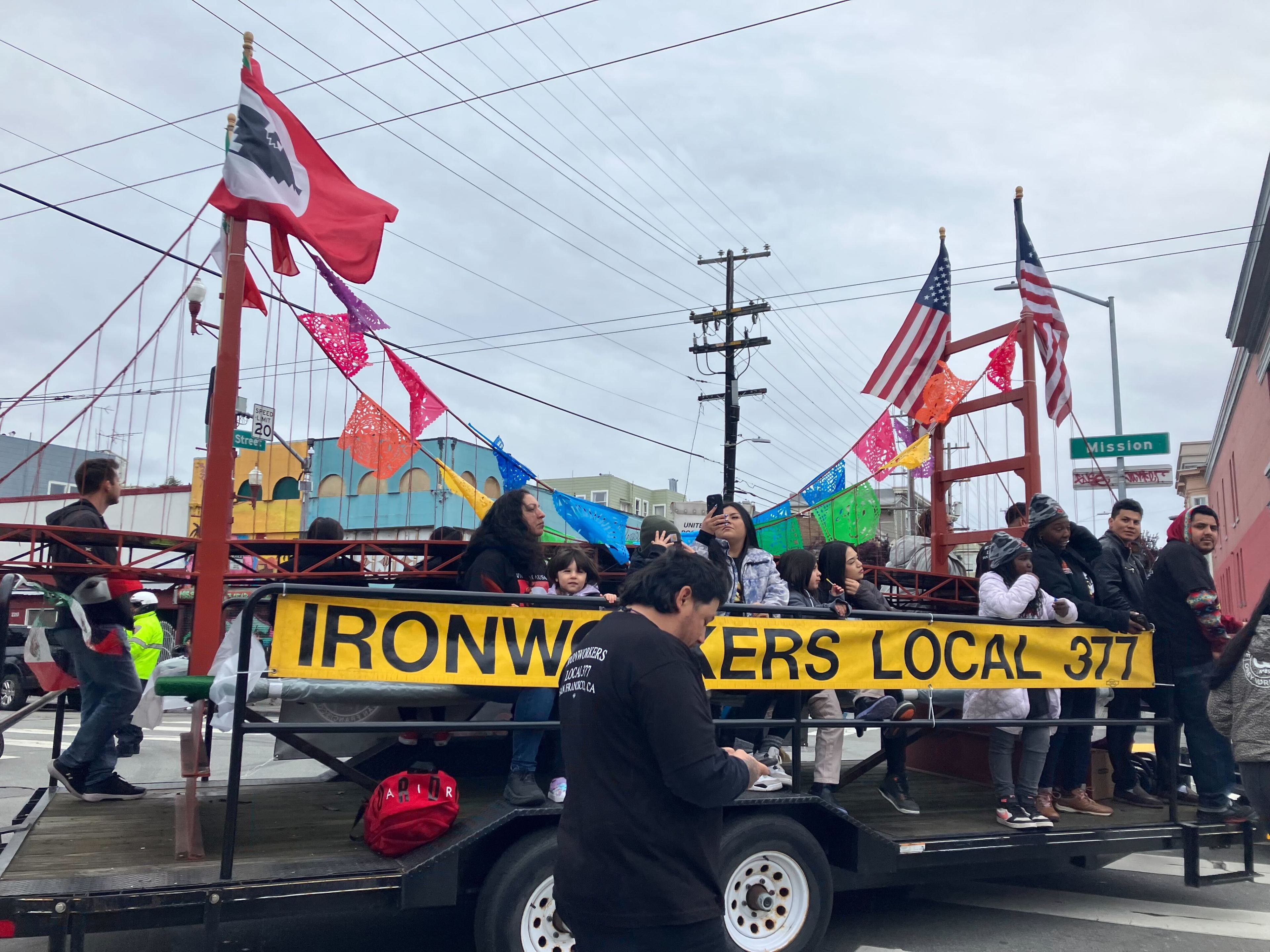 A parade float with people, flags, and a &quot;IRONWORKERS LOCAL 377&quot; banner, moving through a street.