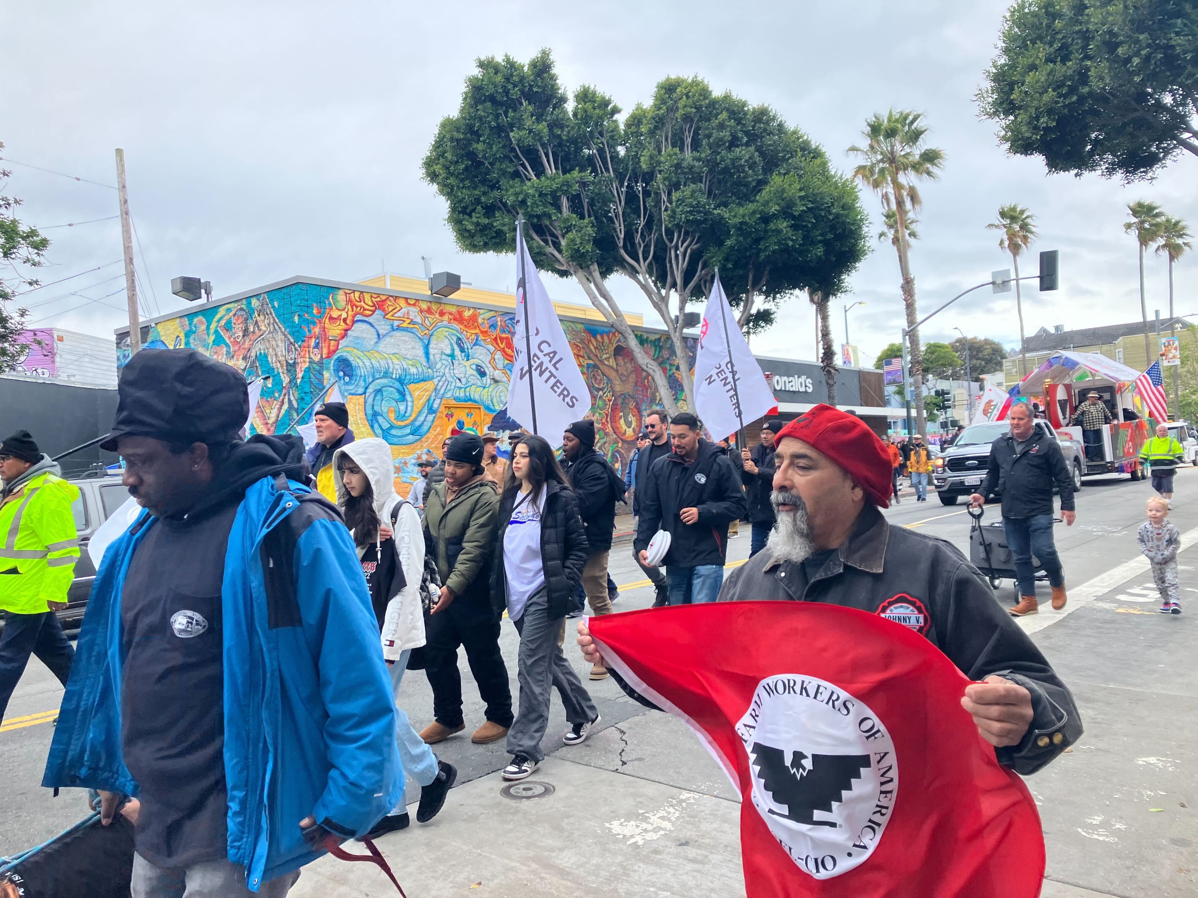 A group of people marching, some carrying banners, with a colorful mural and palm trees in the background.