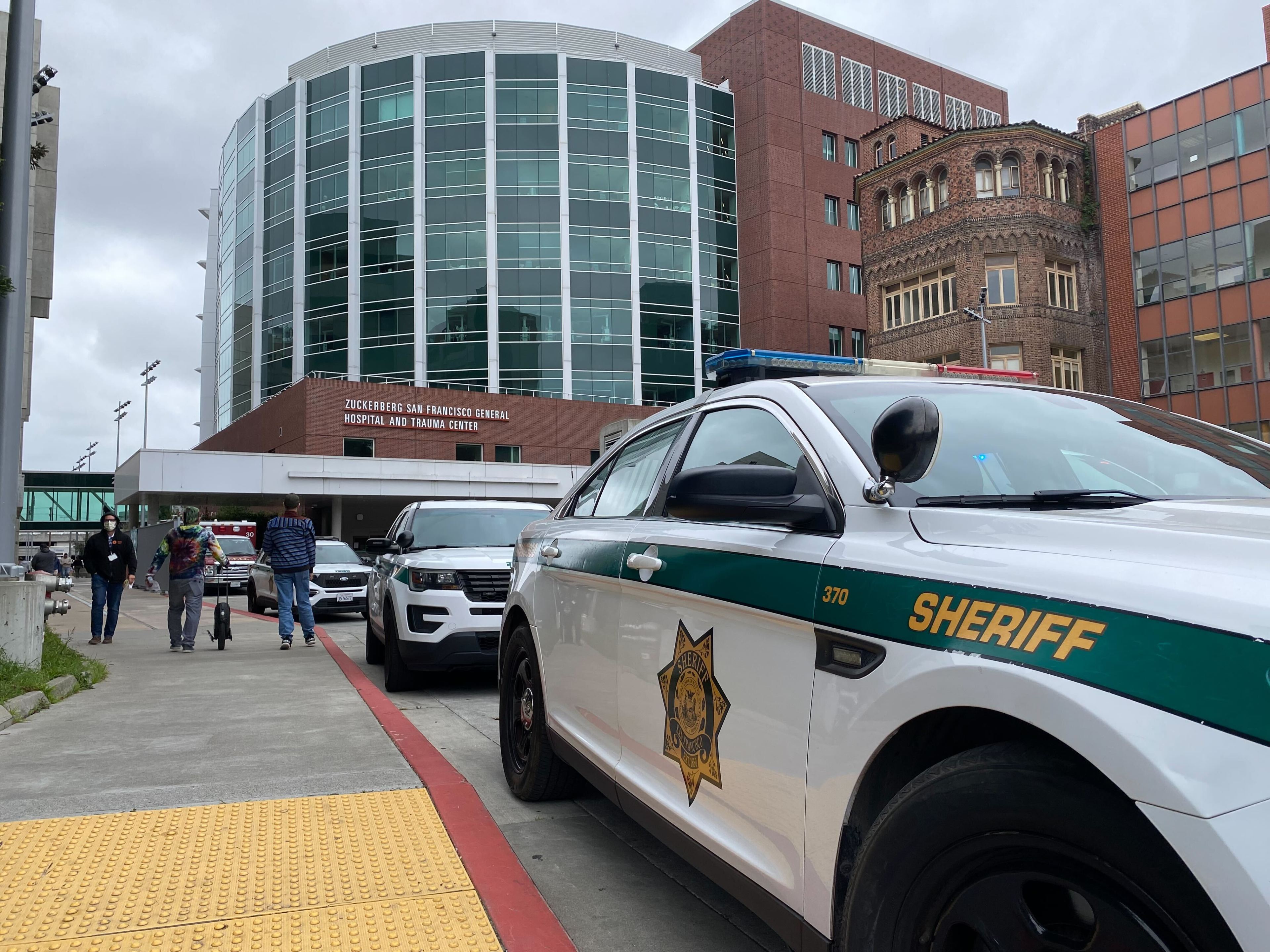 Sheriff cars in front of a hospital with pedestrians nearby.