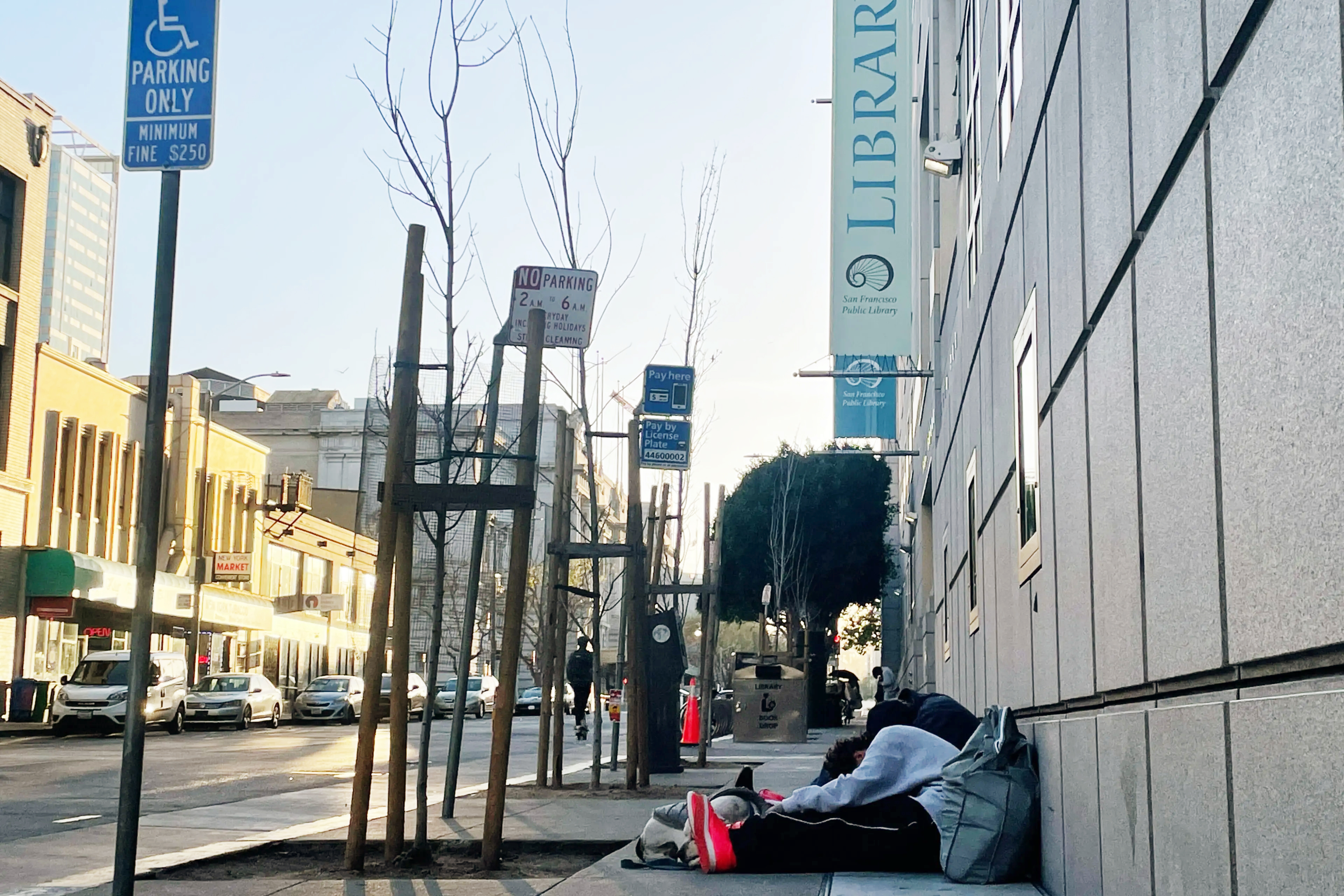 A person lies on a sidewalk next to a building with "LIBRARY" banners, urban setting, daytime.