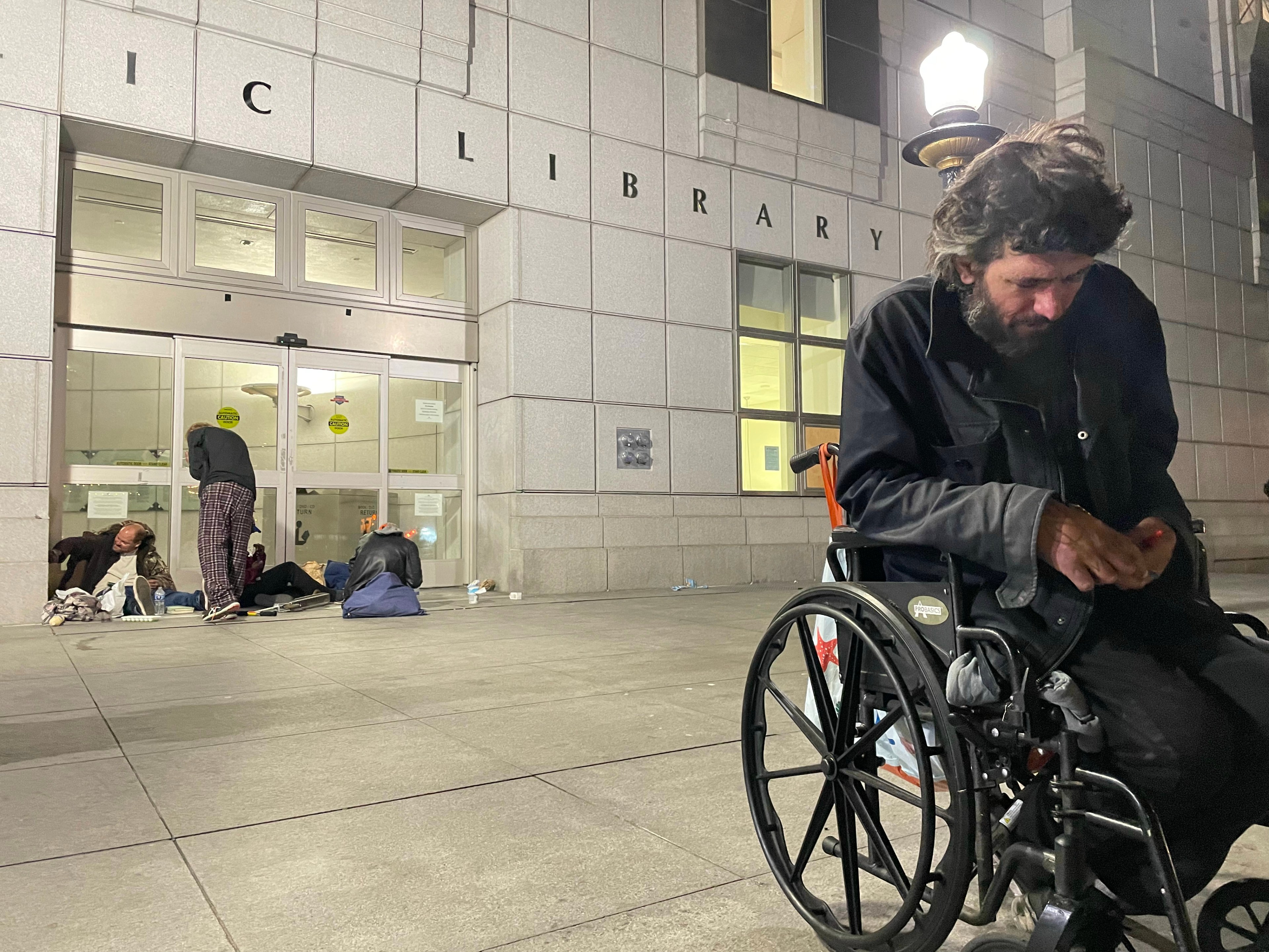 A man in a wheelchair and others sit outside a building labeled &quot;LIBRARY&quot; at night, suggesting urban homelessness.