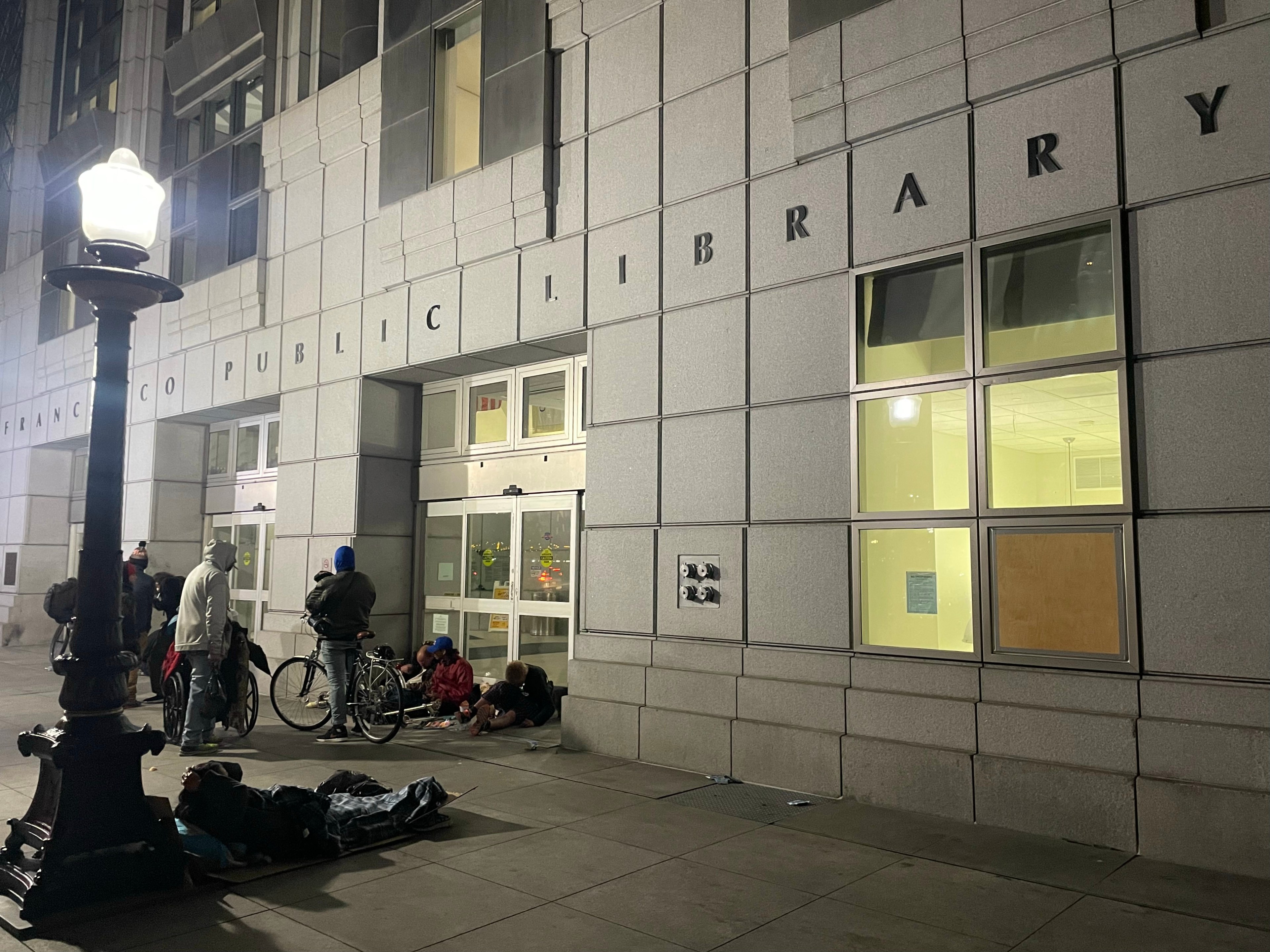 Outside a public library at night, with people gathered by the entrance and others lying on the pavement.