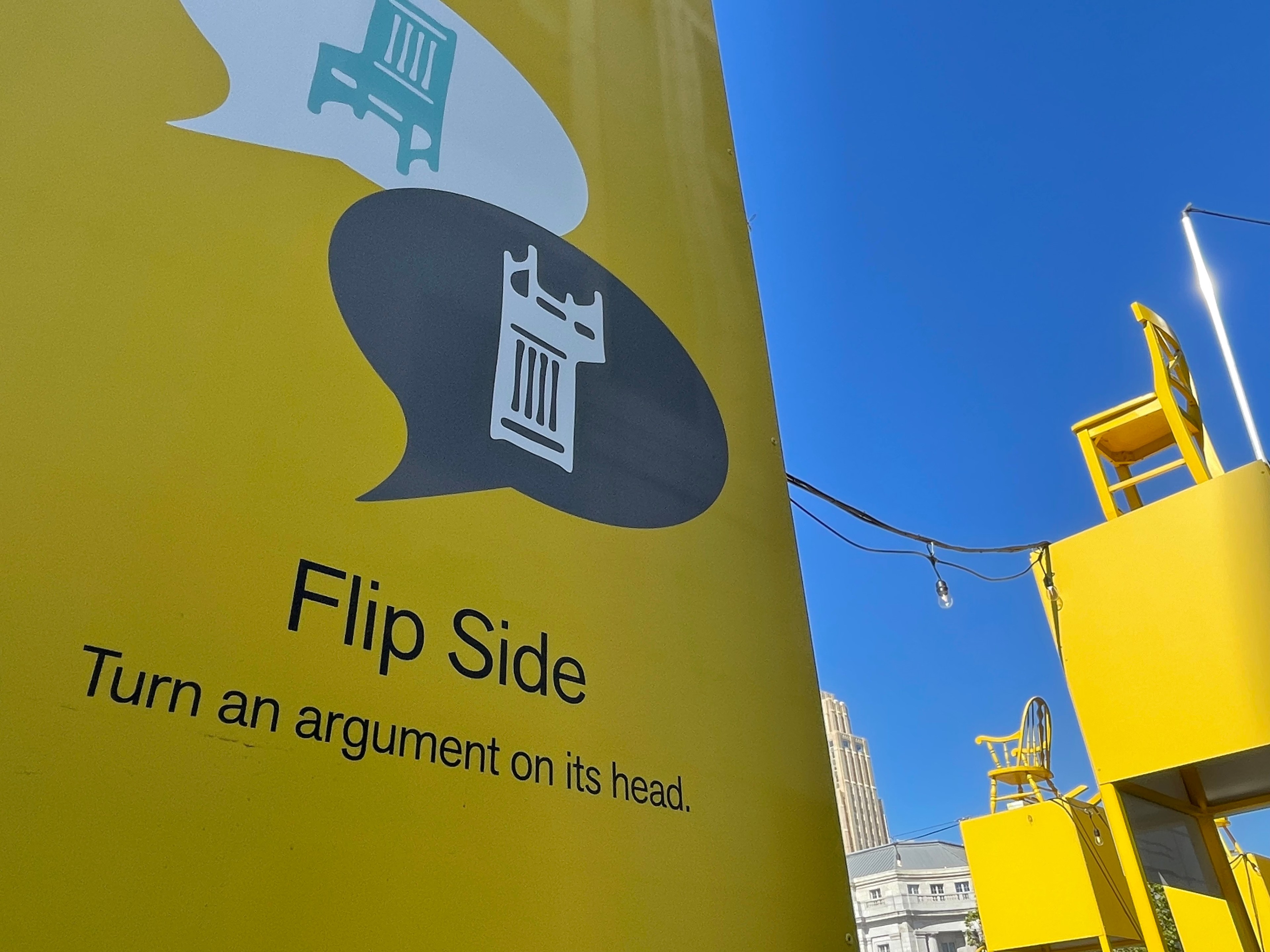 A yellow sign with text &quot;Flip Side Turn an argument on its head,&quot; two inverted speech bubbles with chair icons, under a blue sky.