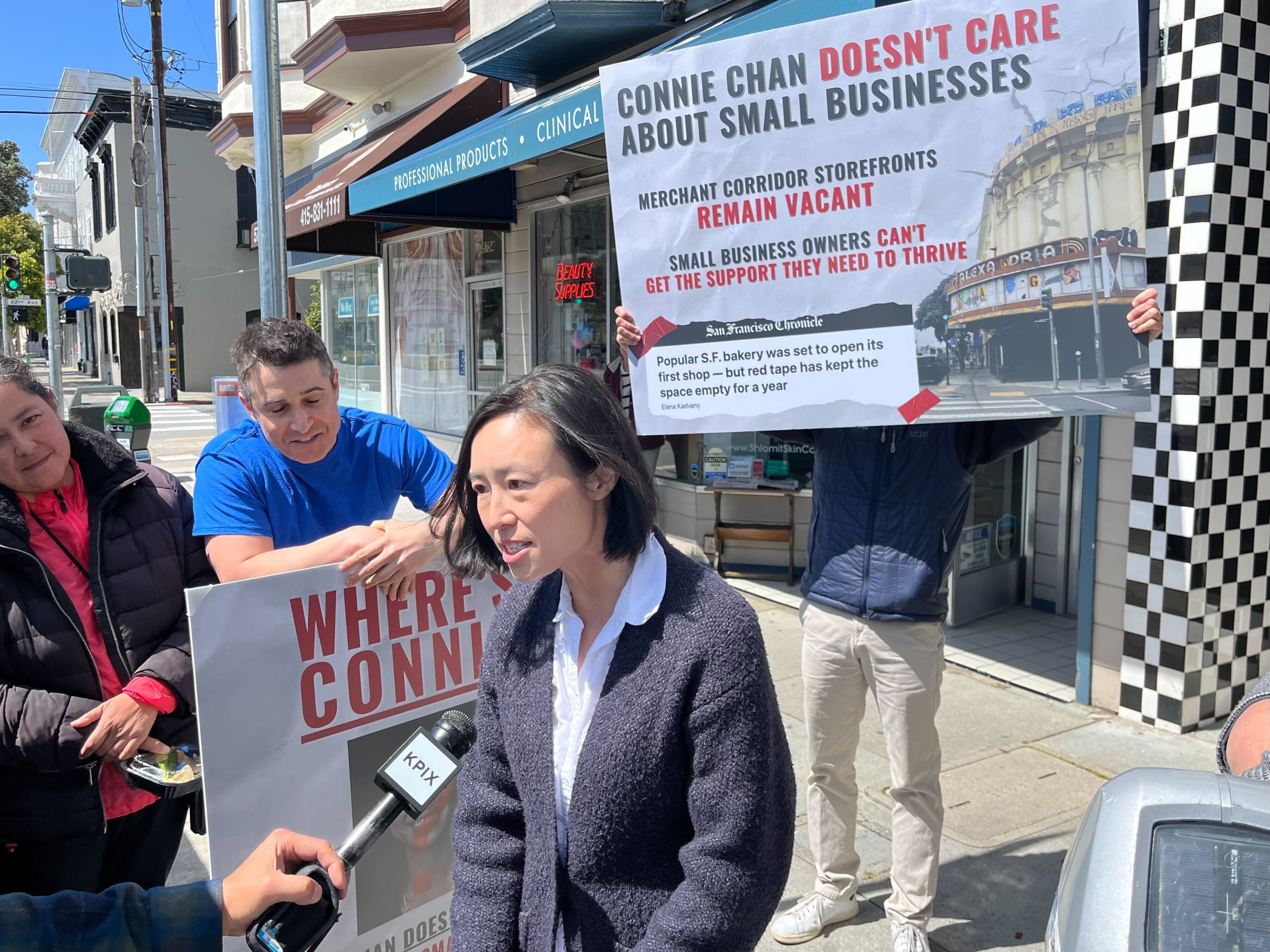 A person is being interviewed on a sunny street, holding a sign about local business concerns, with others holding a larger banner.