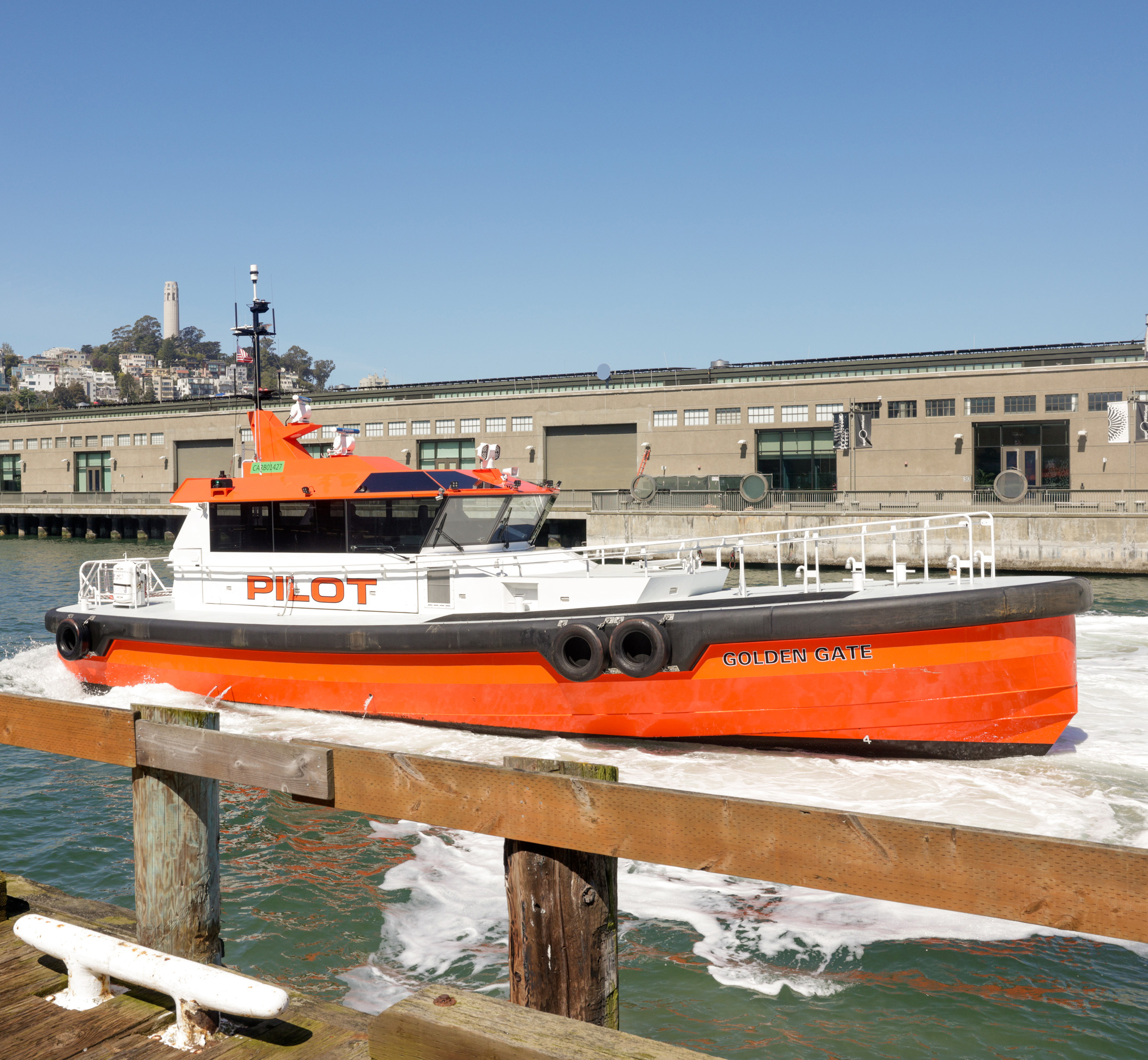An orange and white pilot boat named &quot;GOLDEN GATE&quot; navigates near a pier with a city hill backdrop.
