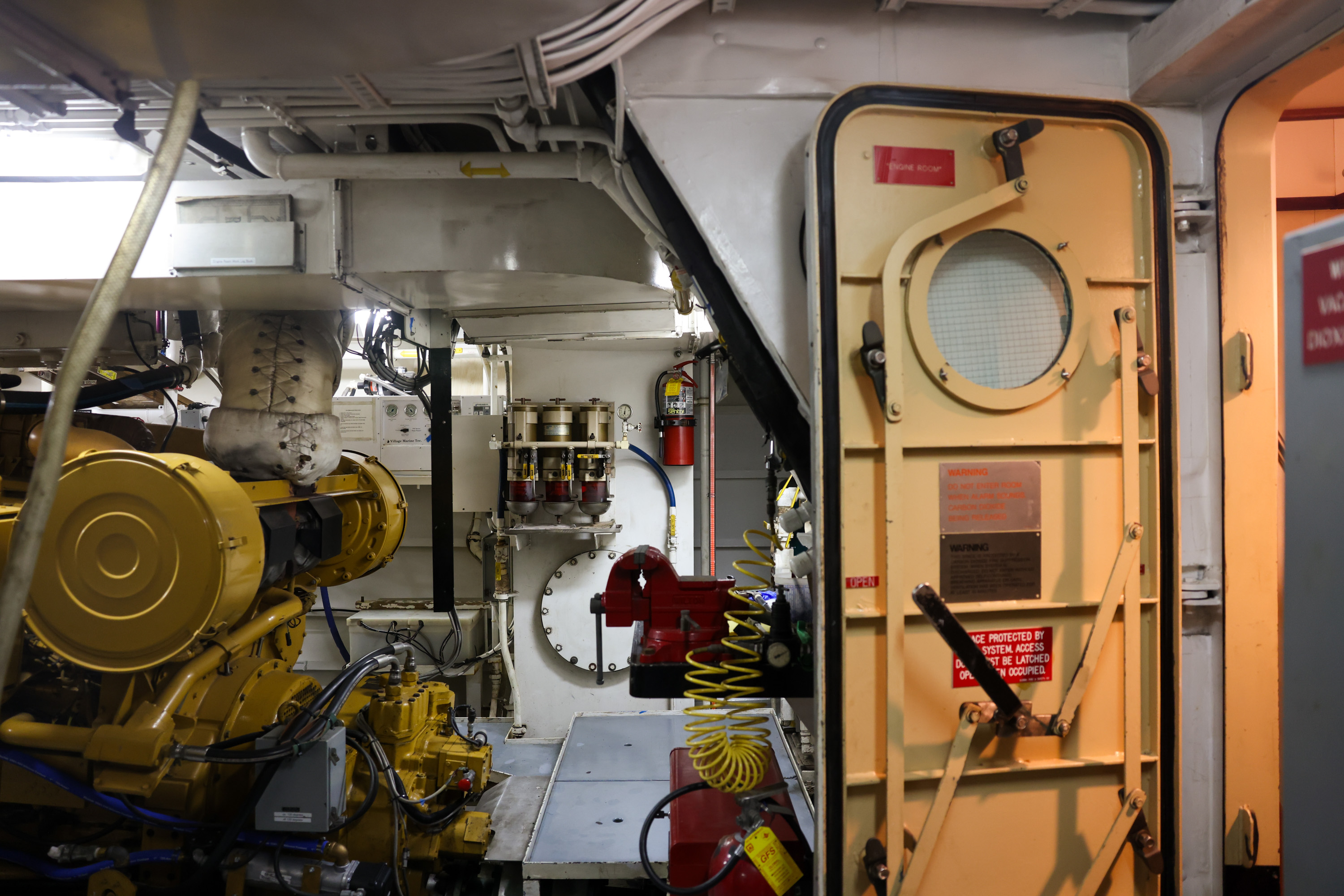 Inside a ship's engine room with machinery, piping, and a door with a porthole.