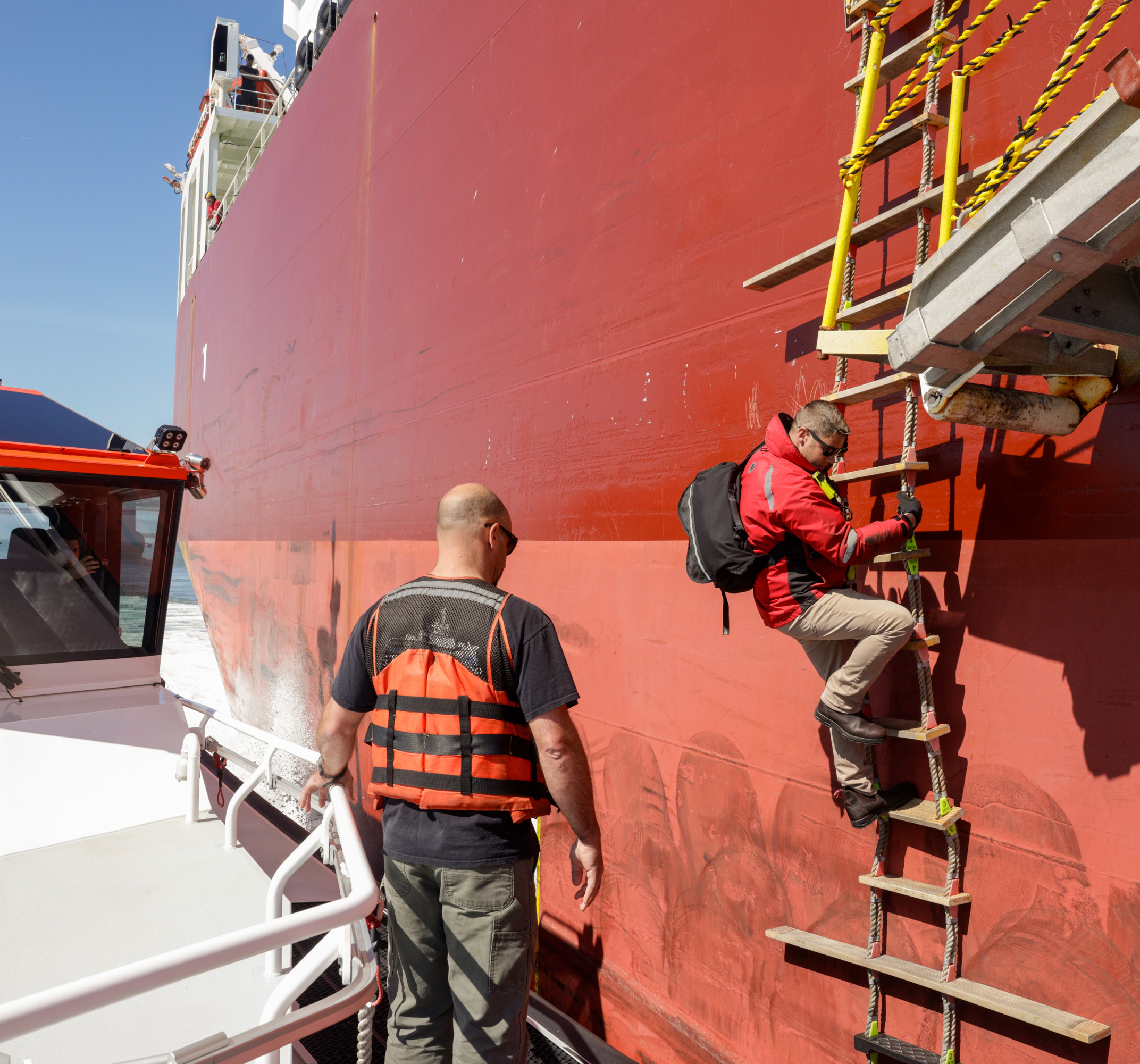 A person in a red jacket climbs a rope ladder onto a large red ship; another person watches from a smaller vessel.