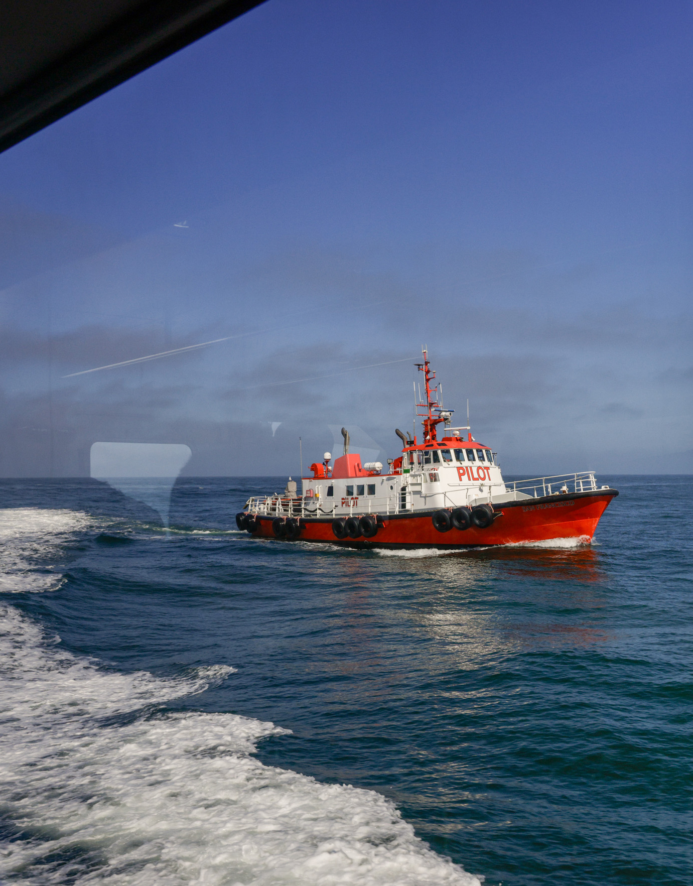 A pilot boat at sea, viewed from a window, leaving a wake under a blue sky.