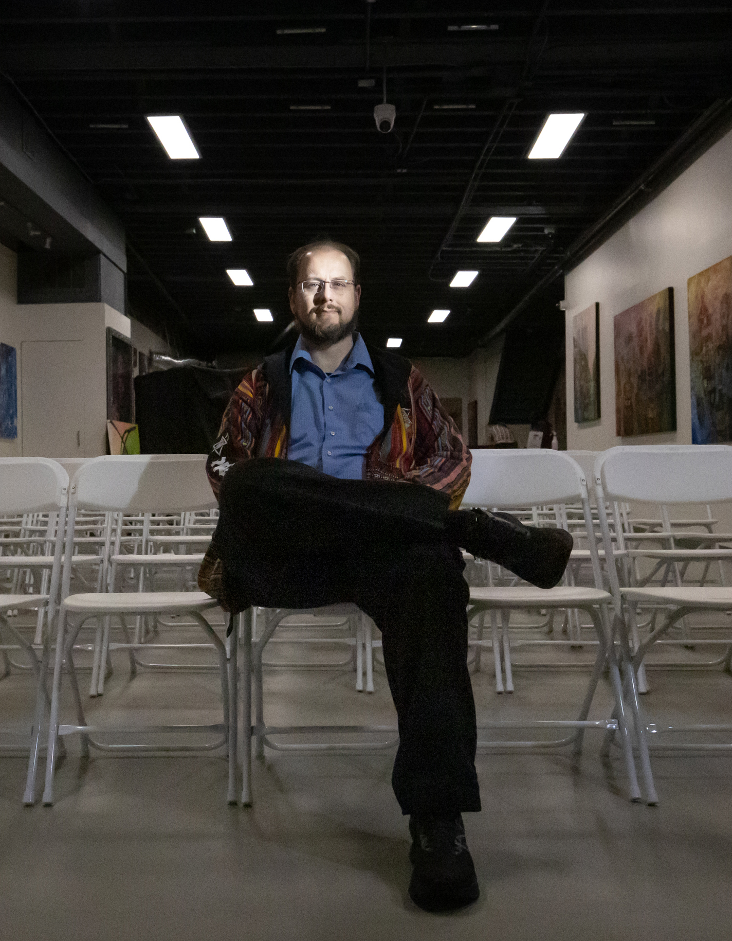 A man is sitting cross-legged on a chair in an art gallery with paintings on the walls and rows of white chairs.