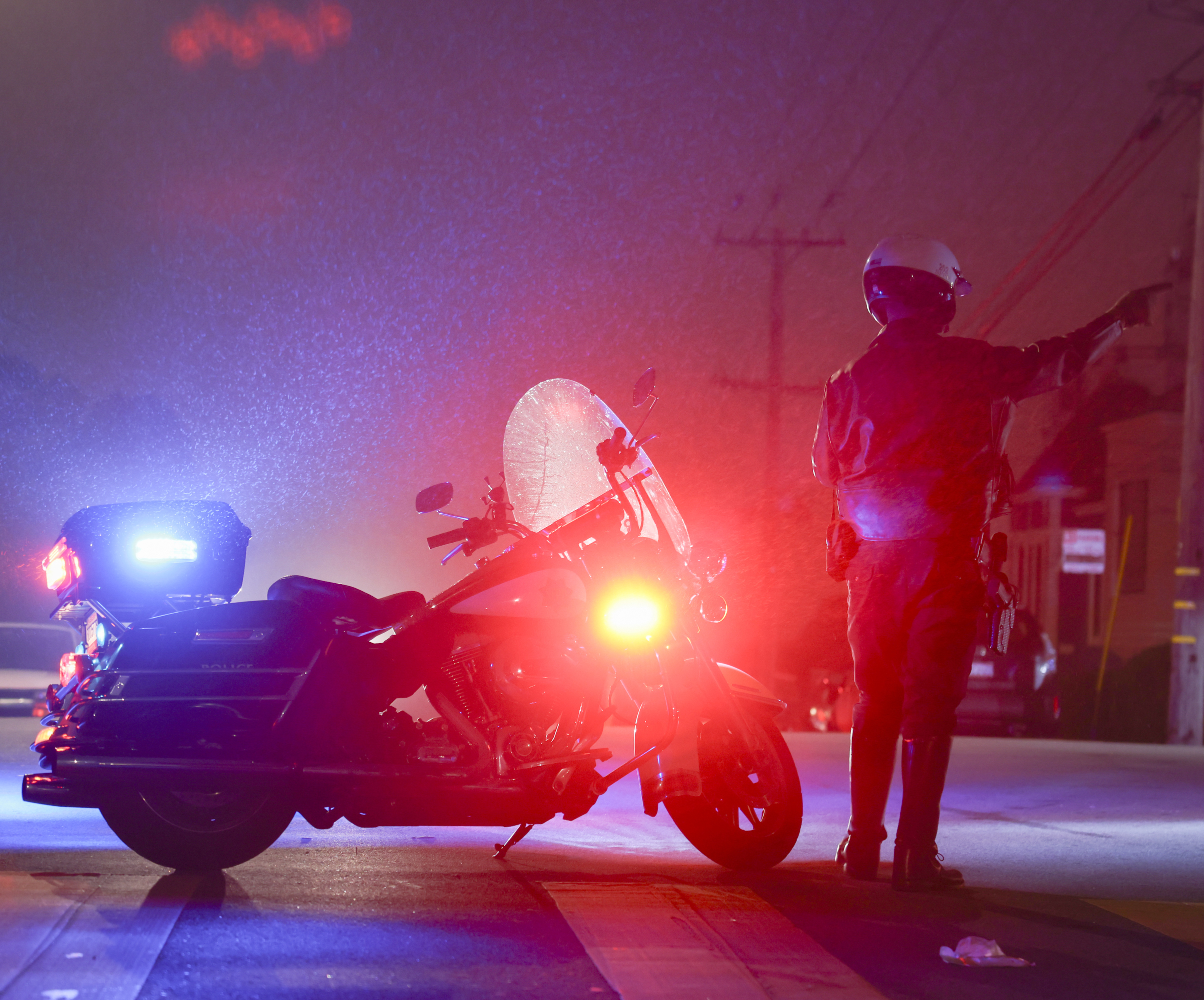 A police officer beside their motorcycle at night, pointing ahead, with emergency lights on.