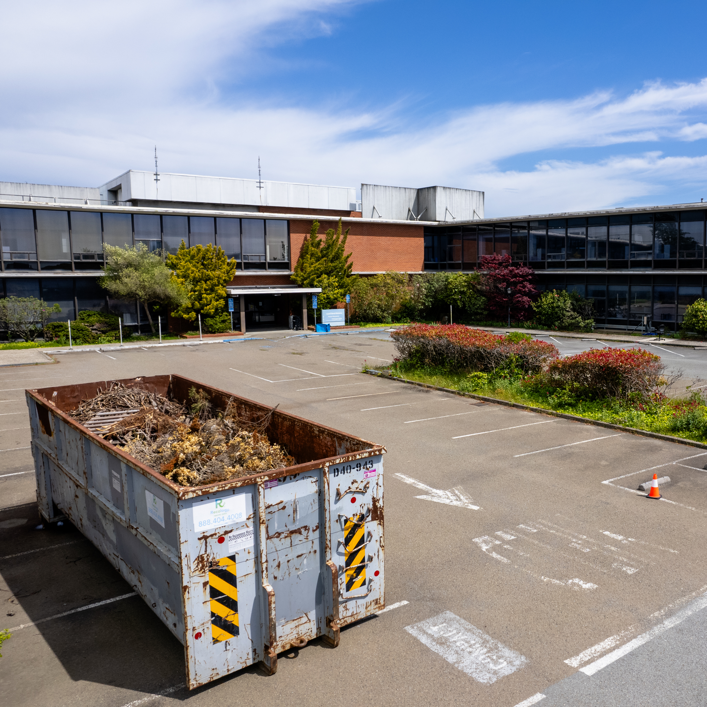 An overgrown parking lot with a rusty dumpster, surrounded by a deserted building under a clear sky.
