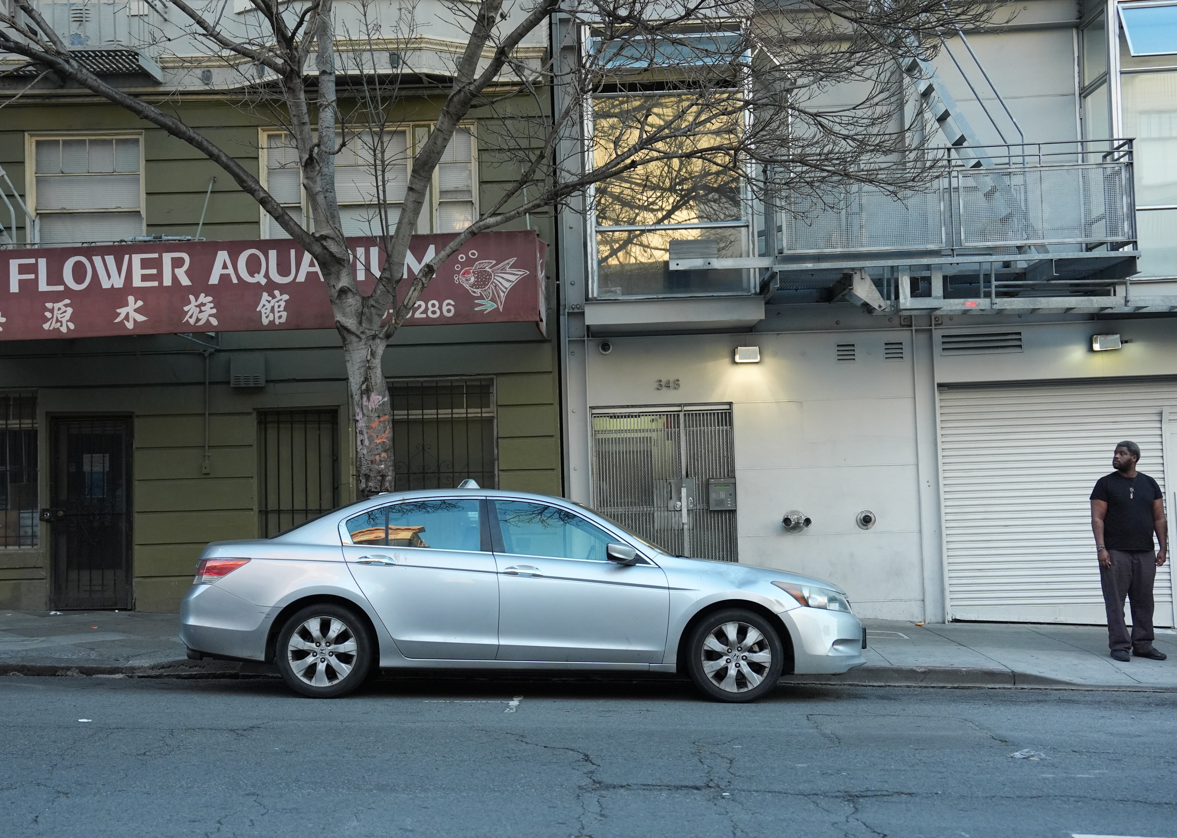 A silver car is parked on a street in front of a building with a &quot;Flower Aquarium&quot; sign; a man stands on the sidewalk to the right.