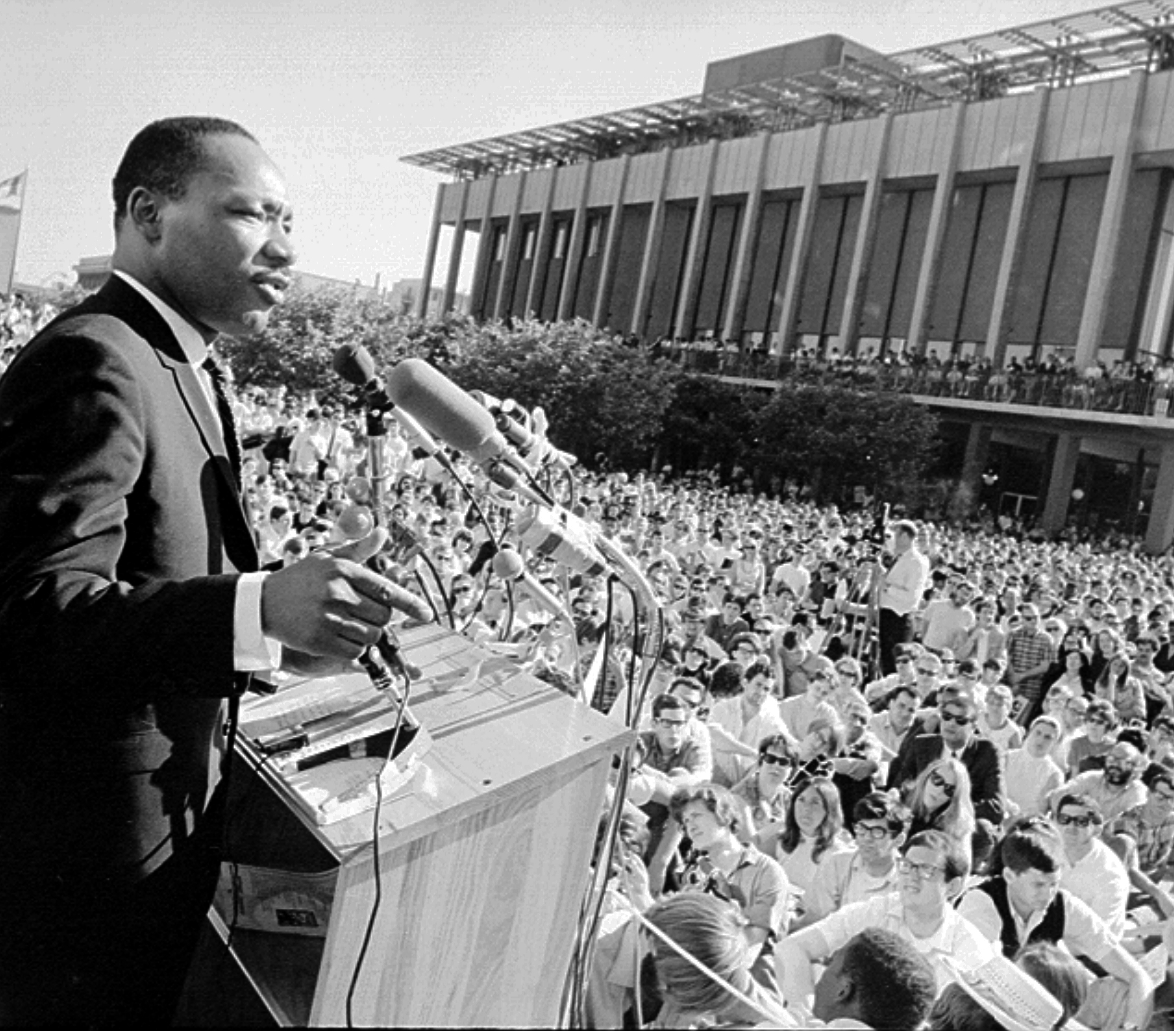 Martin Luther King, Jr. speaks at a podium before a large crowd, outdoors, in a black and white photo.