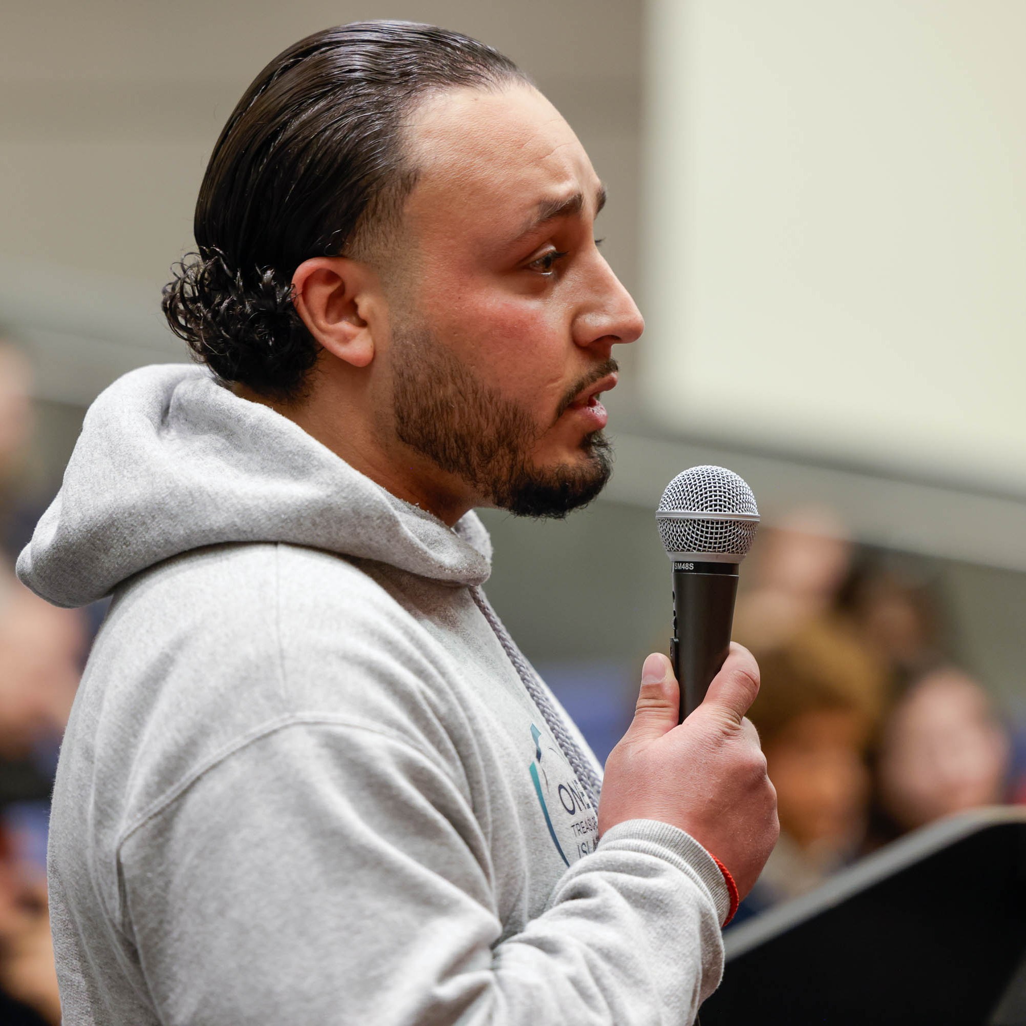 A man with long black hair and a gray hoodie speaks at a podium, seen in a profile side-angle.
