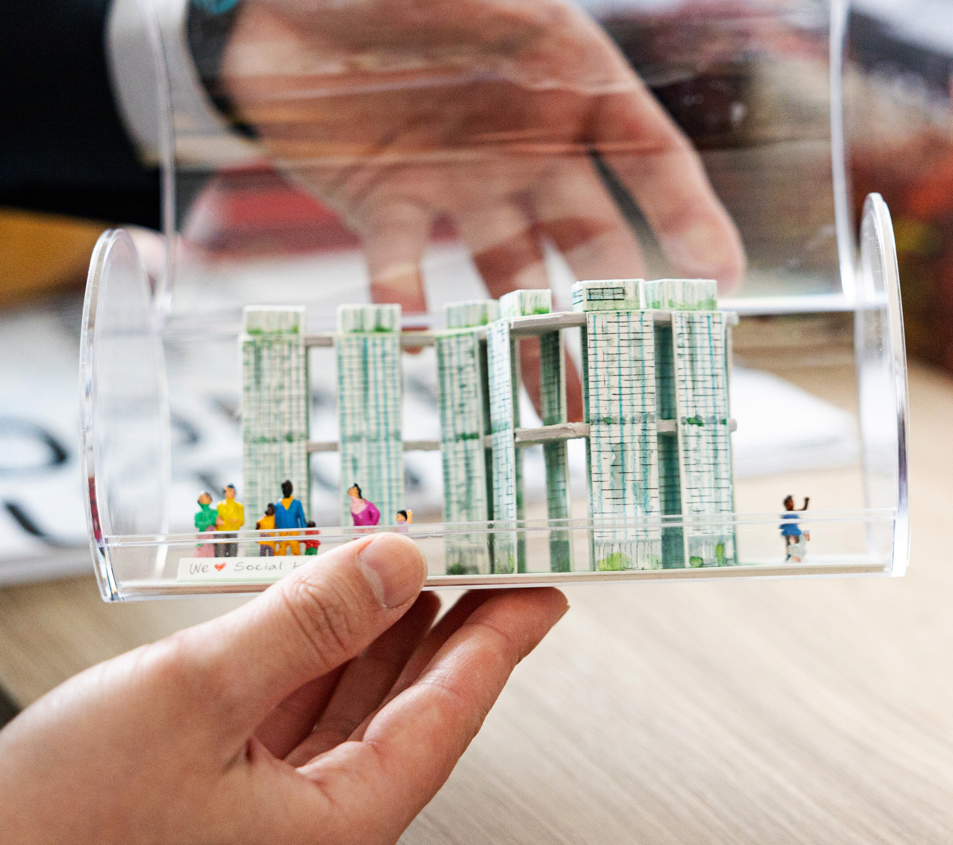A hand holds a clear model with miniature people and green high-rise structures inside.