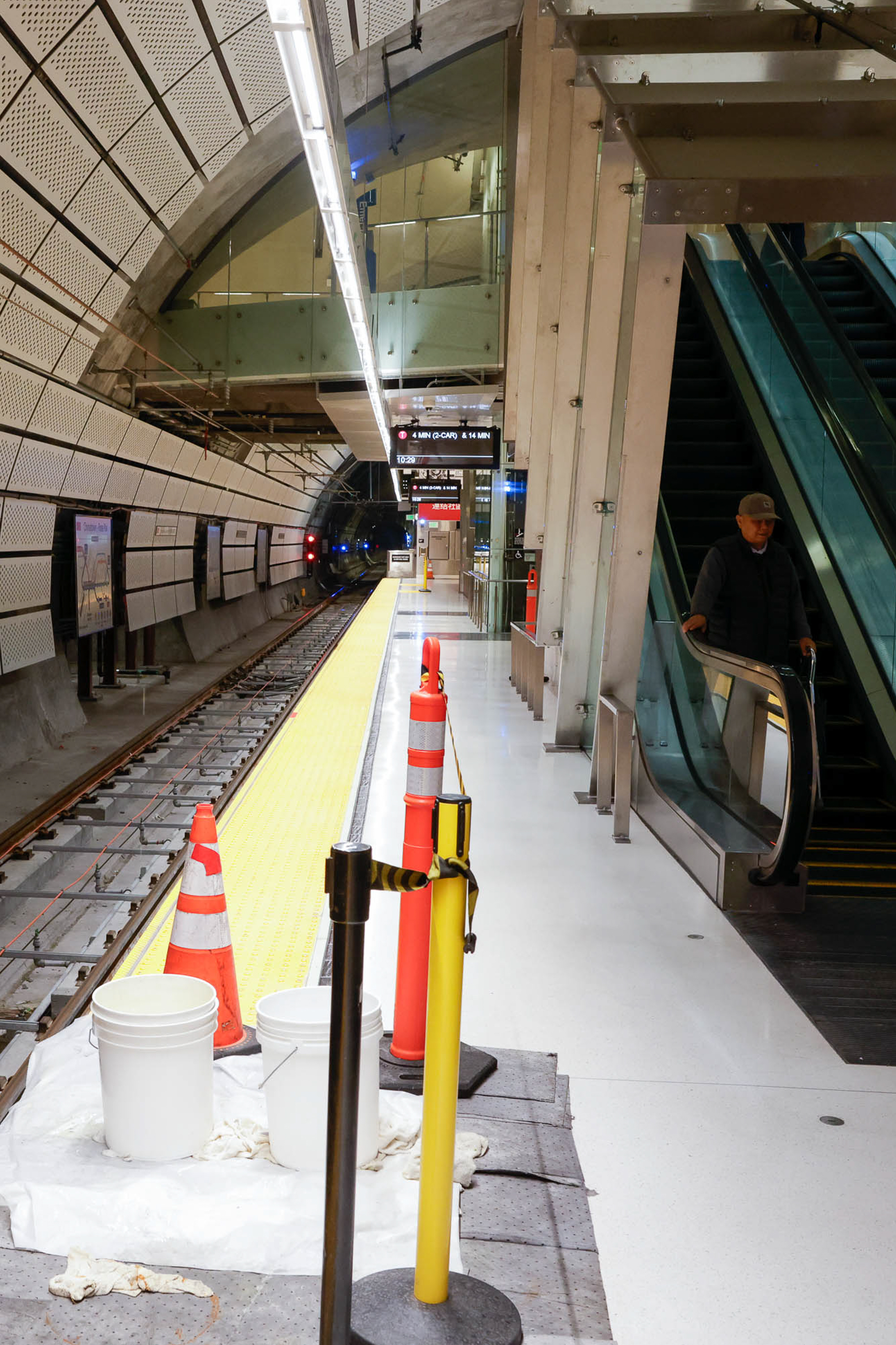 An empty subway station platform with a man riding an escalator, construction cones with buckets collecting dripping water.