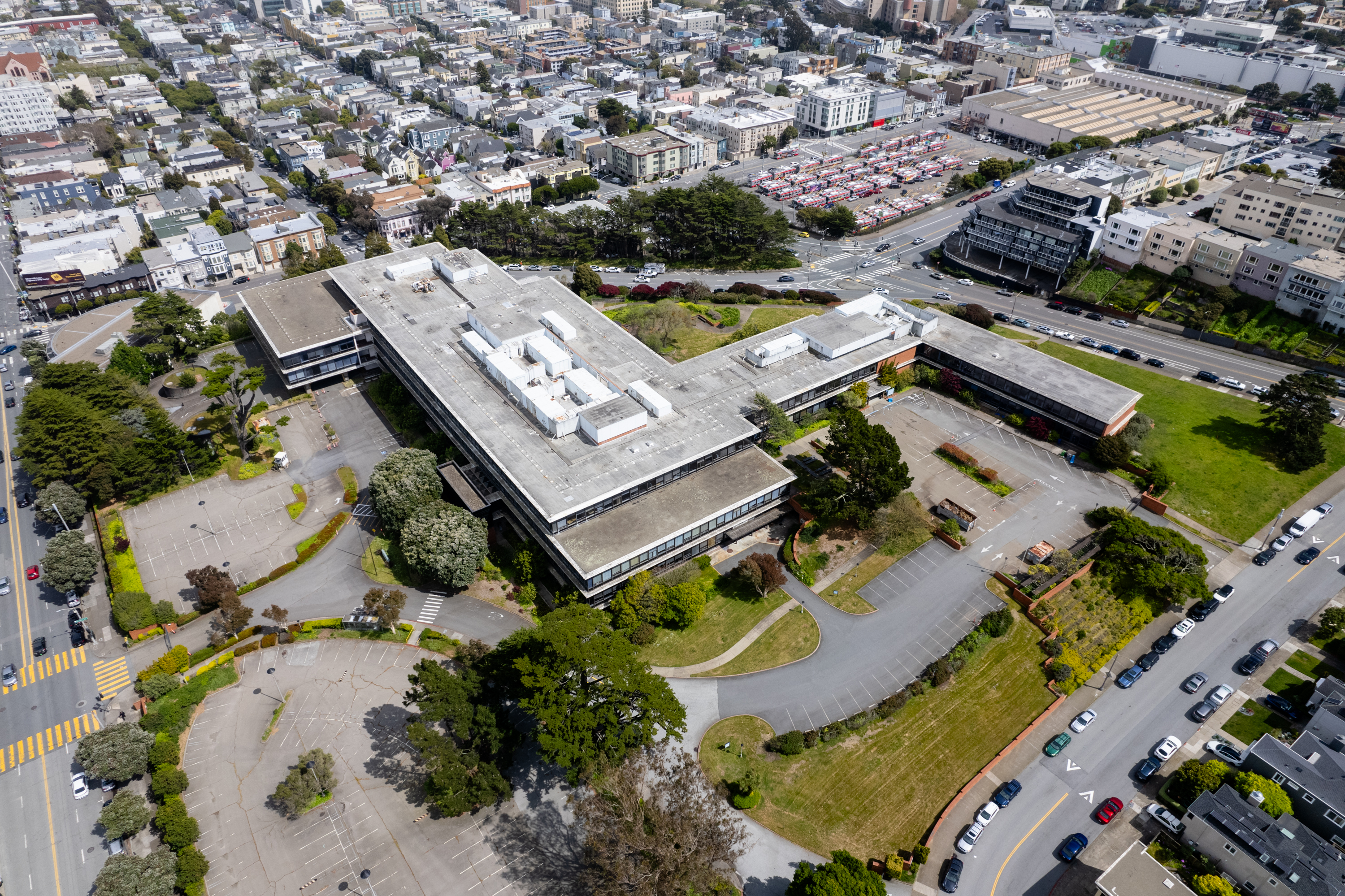 An aerial view of a large, flat-roofed building with parking lot, surrounded by urban streets and densely packed houses.