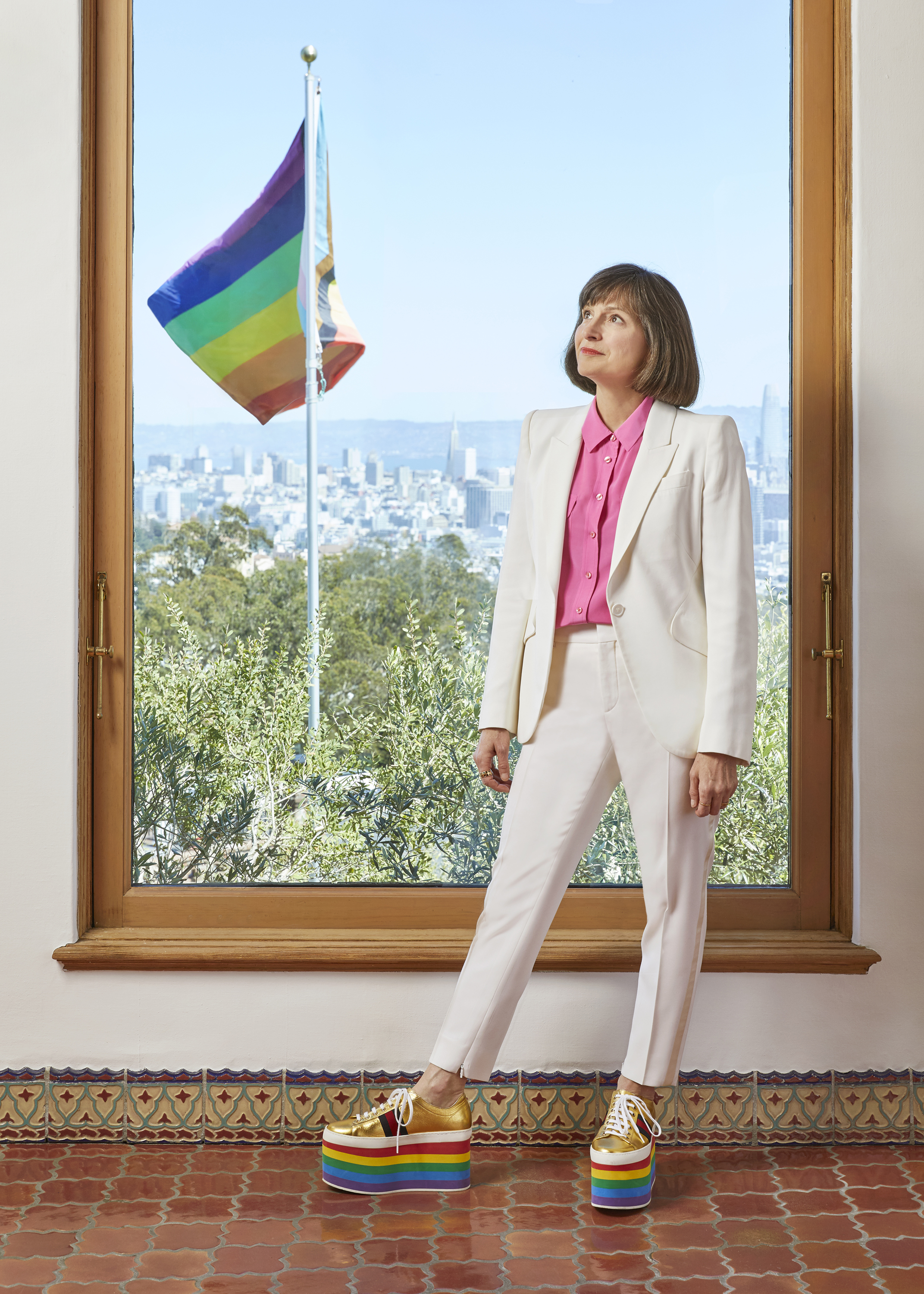 A person stands by a window, wearing a white suit and rainbow-platform sneakers, with a pride flag outside.