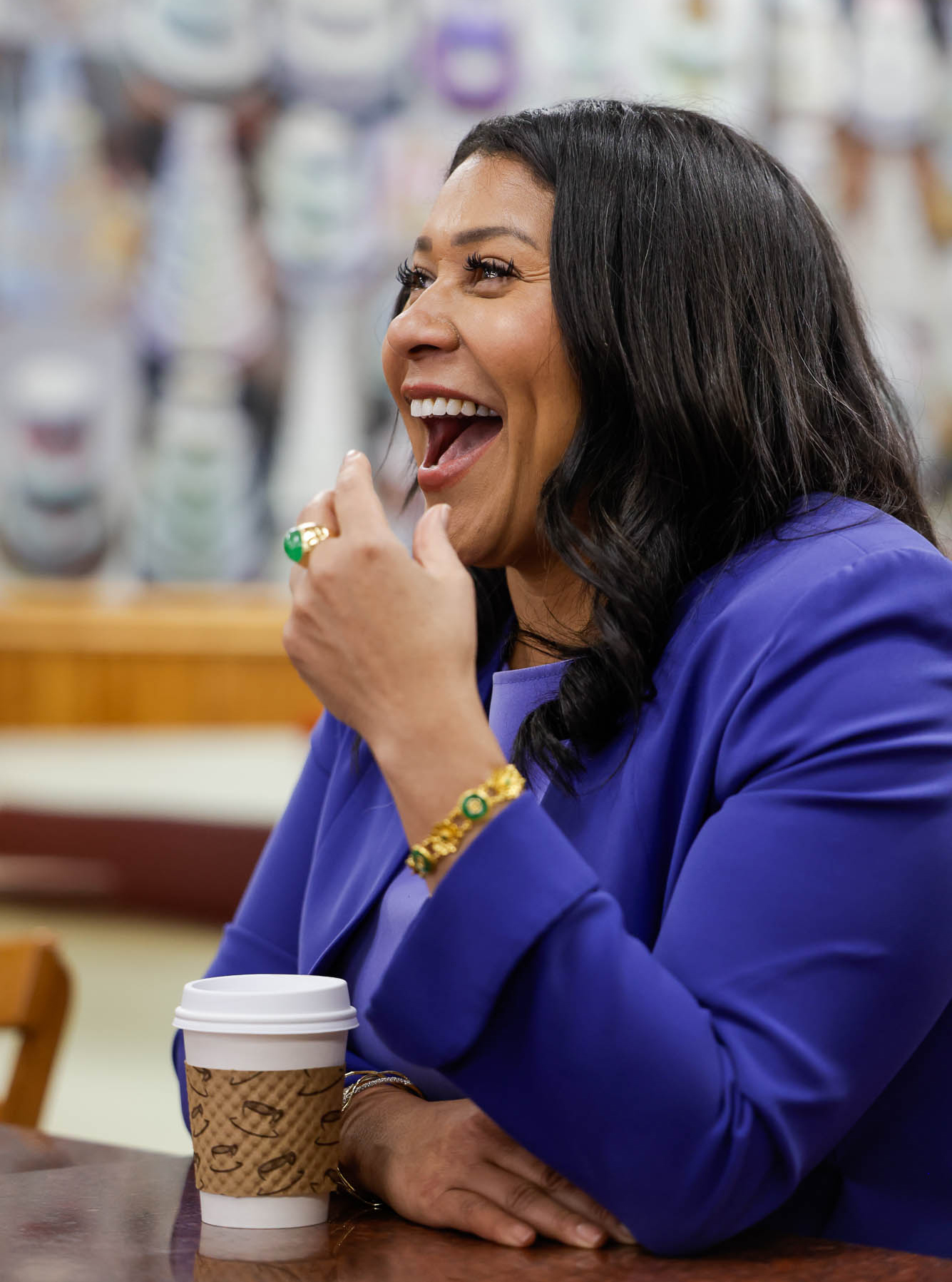 A woman in a blue blazer is laughing, with a coffee cup on the table. She's wearing jewelry.