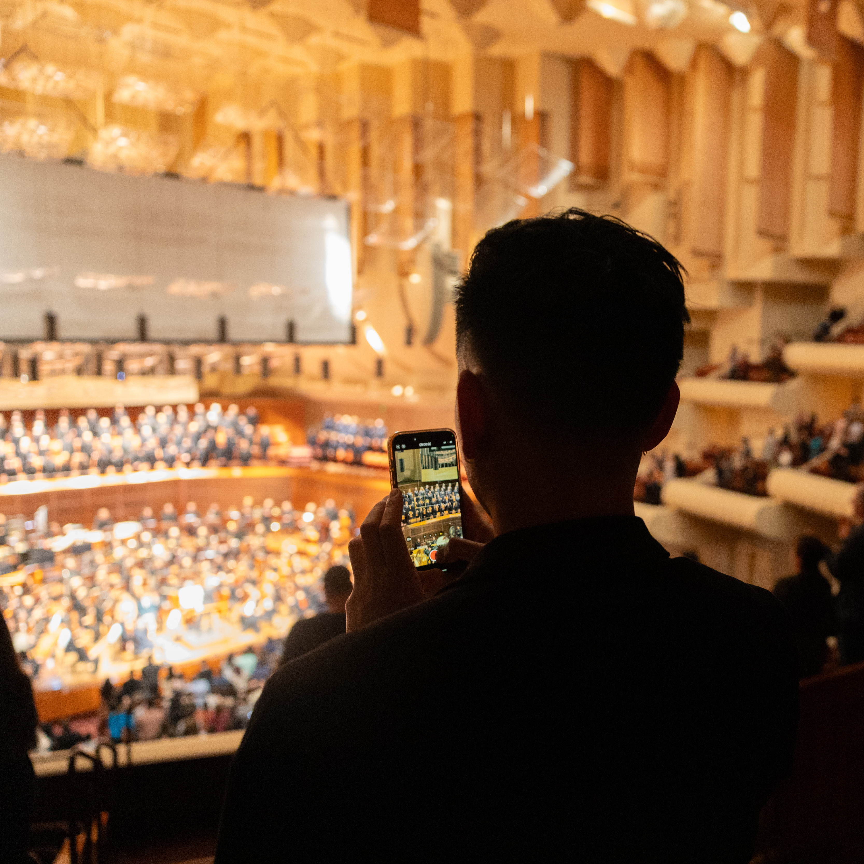 A person is taking a photo of an orchestra in a concert hall with their smartphone.