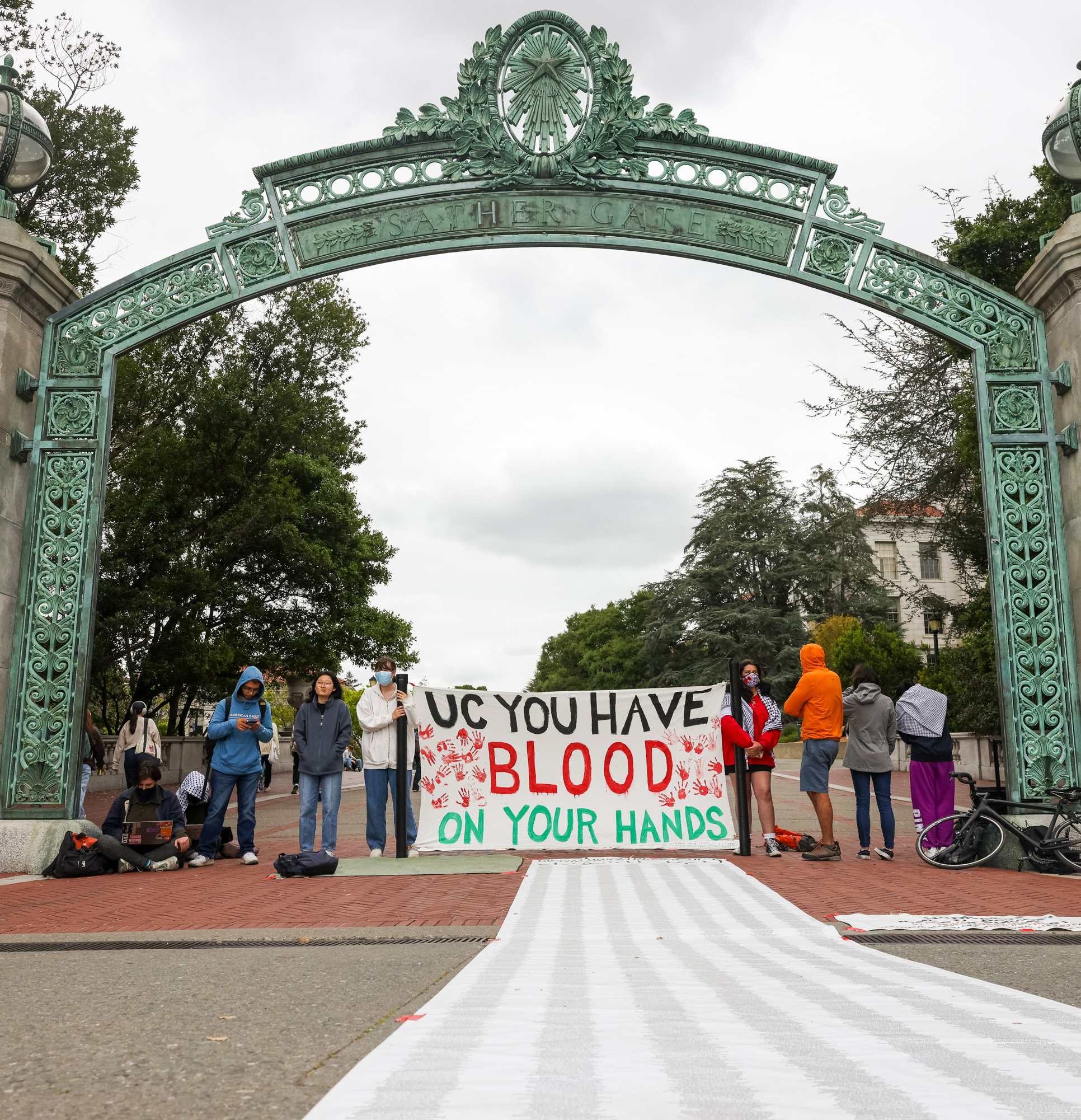A group of people stands under an ornate archway, holding a sign that says &quot;UC YOU HAVE BLOOD ON YOUR HANDS.&quot;