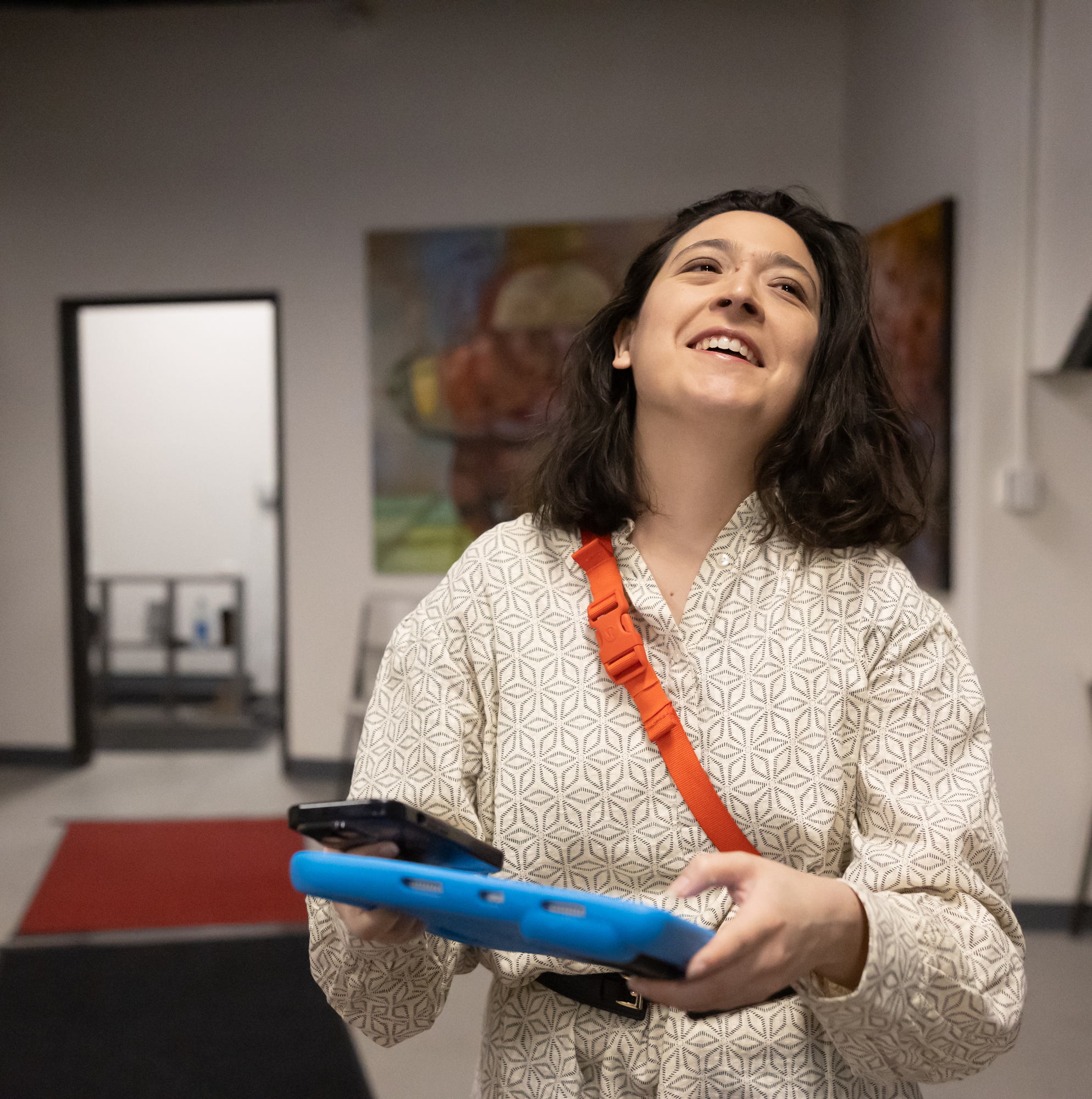 A smiling woman holds an iPad and stands in an indoor setting with artwork on the walls.