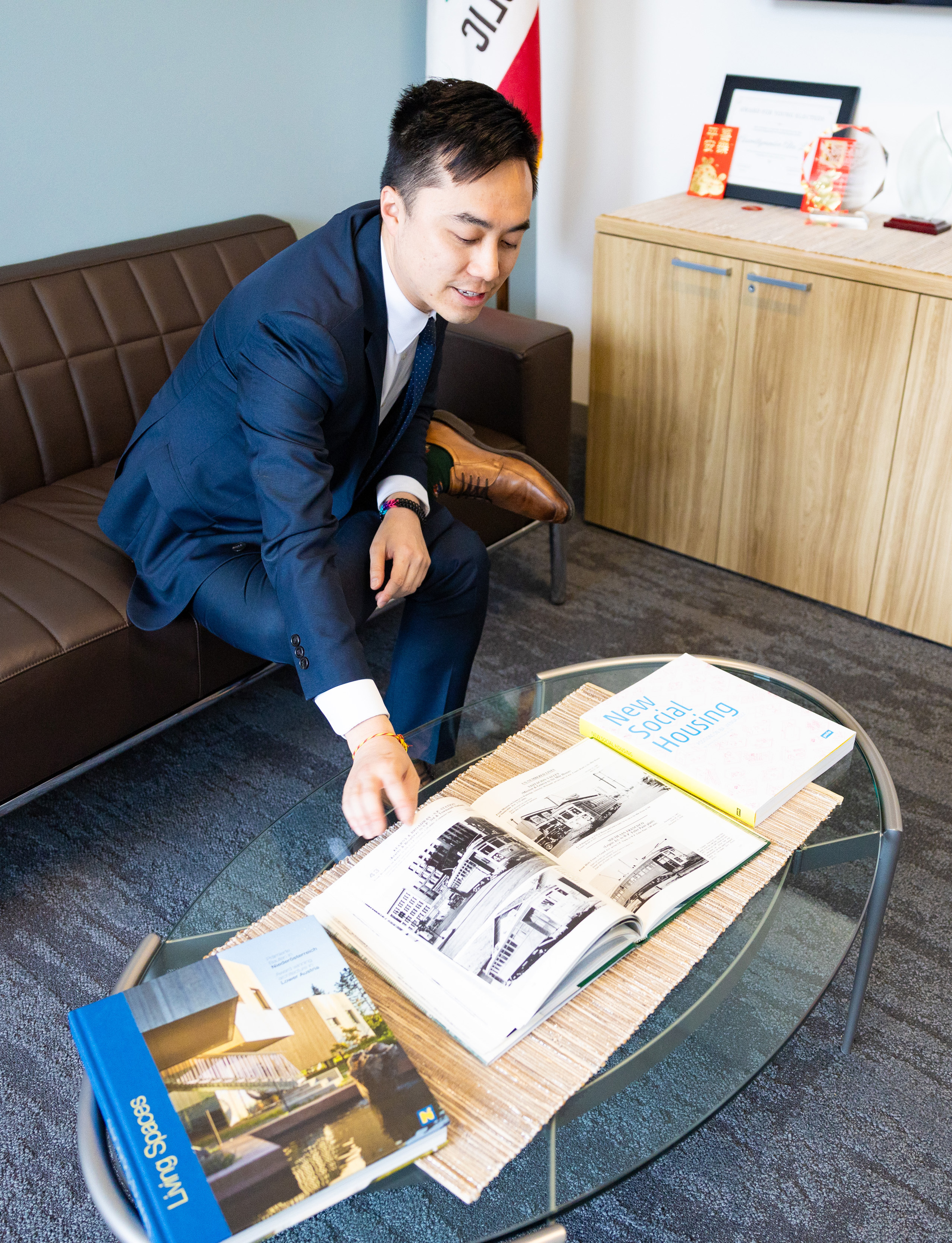A man in a suit leans over a glass table, examining open magazines in an office setting.
