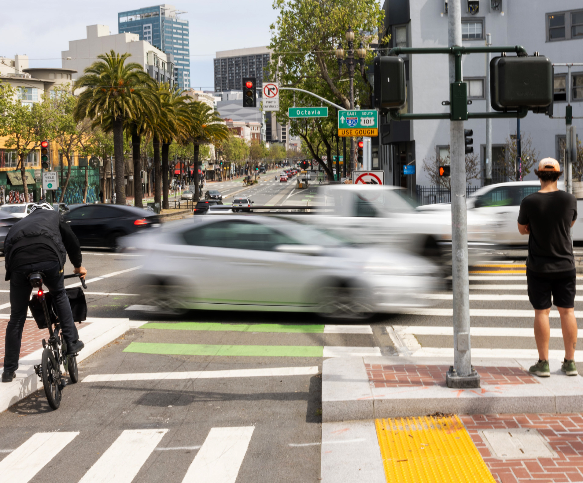 Cyclist and pedestrian waiting at a busy city intersection with blurred moving cars and traffic lights.