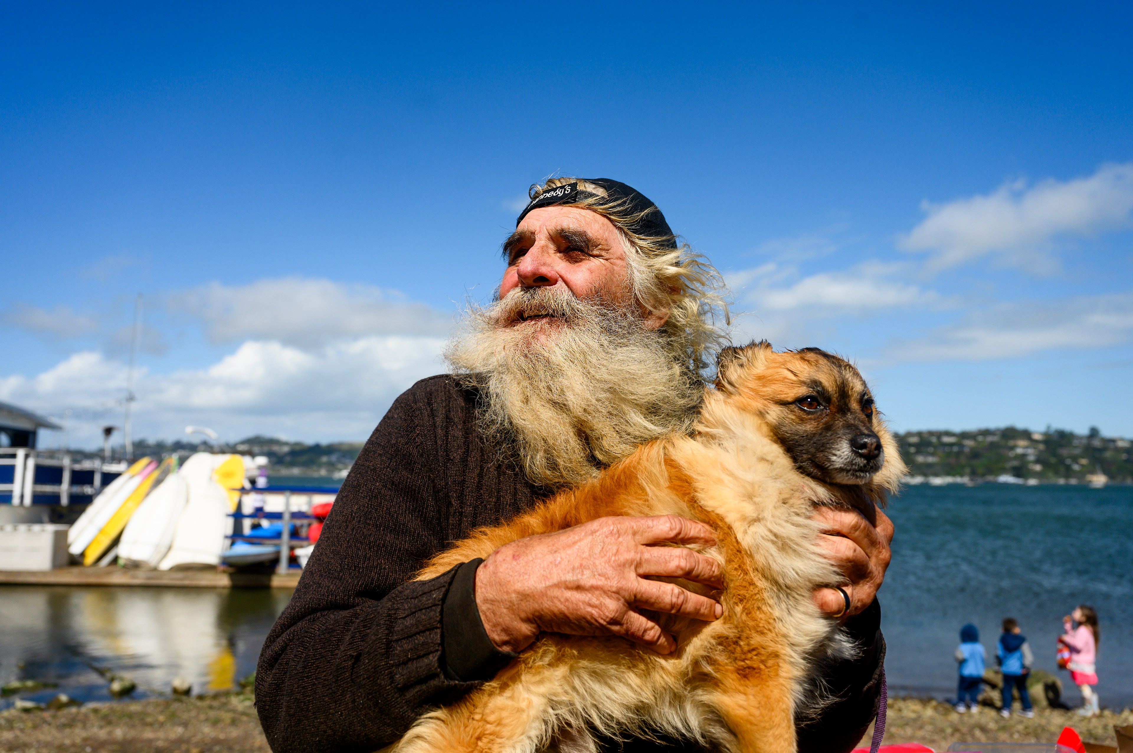 A man with a long beard holds a dog by the water, with children and boats in the background.