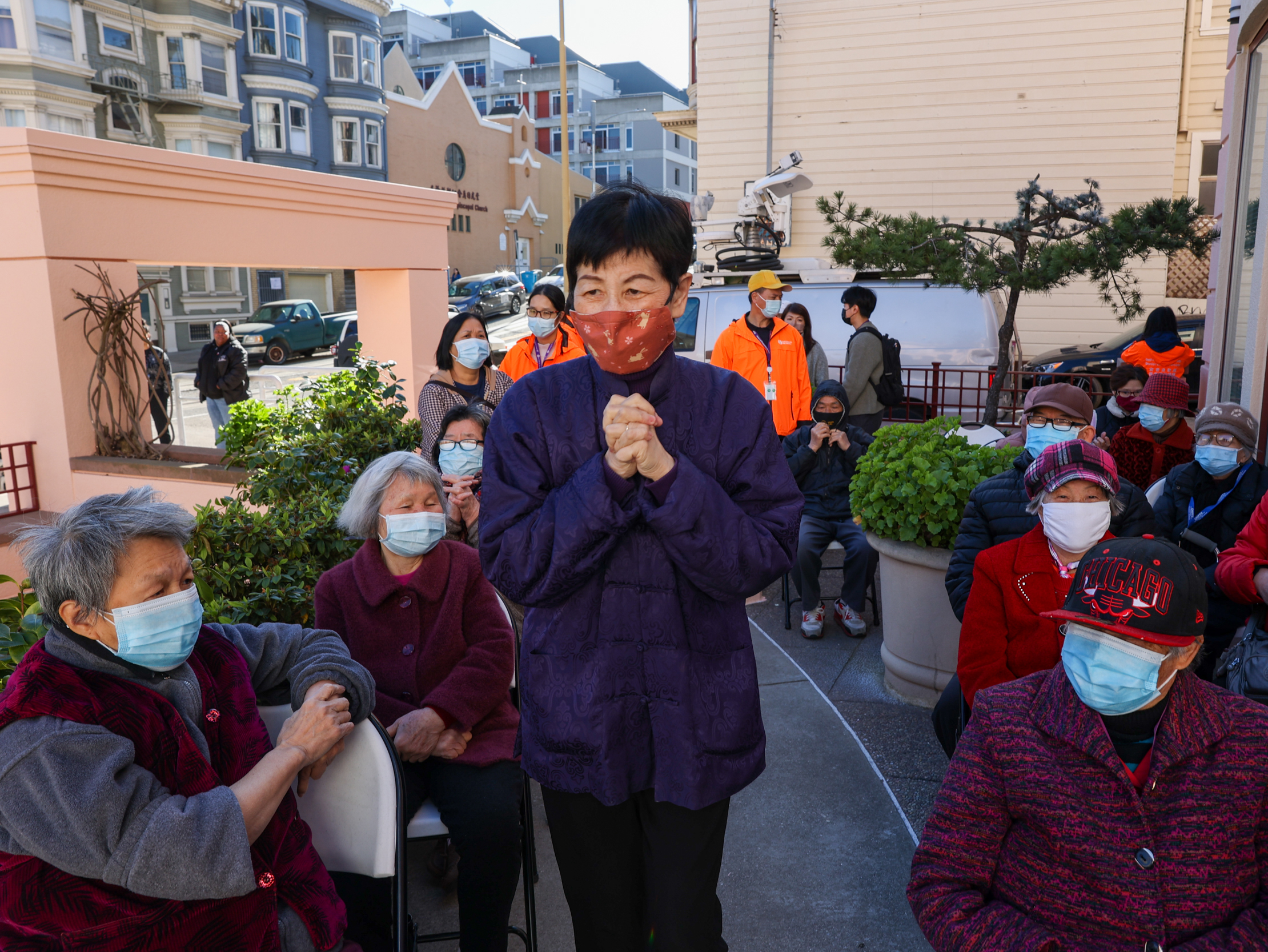 A group of masked seniors gather outdoors; a central figure in purple seems to be speaking or gesturing. 