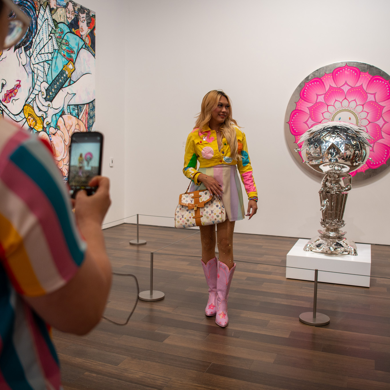 A person is photographed in a colorful outfit inside an art gallery with vibrant artwork and a shiny sculpture.