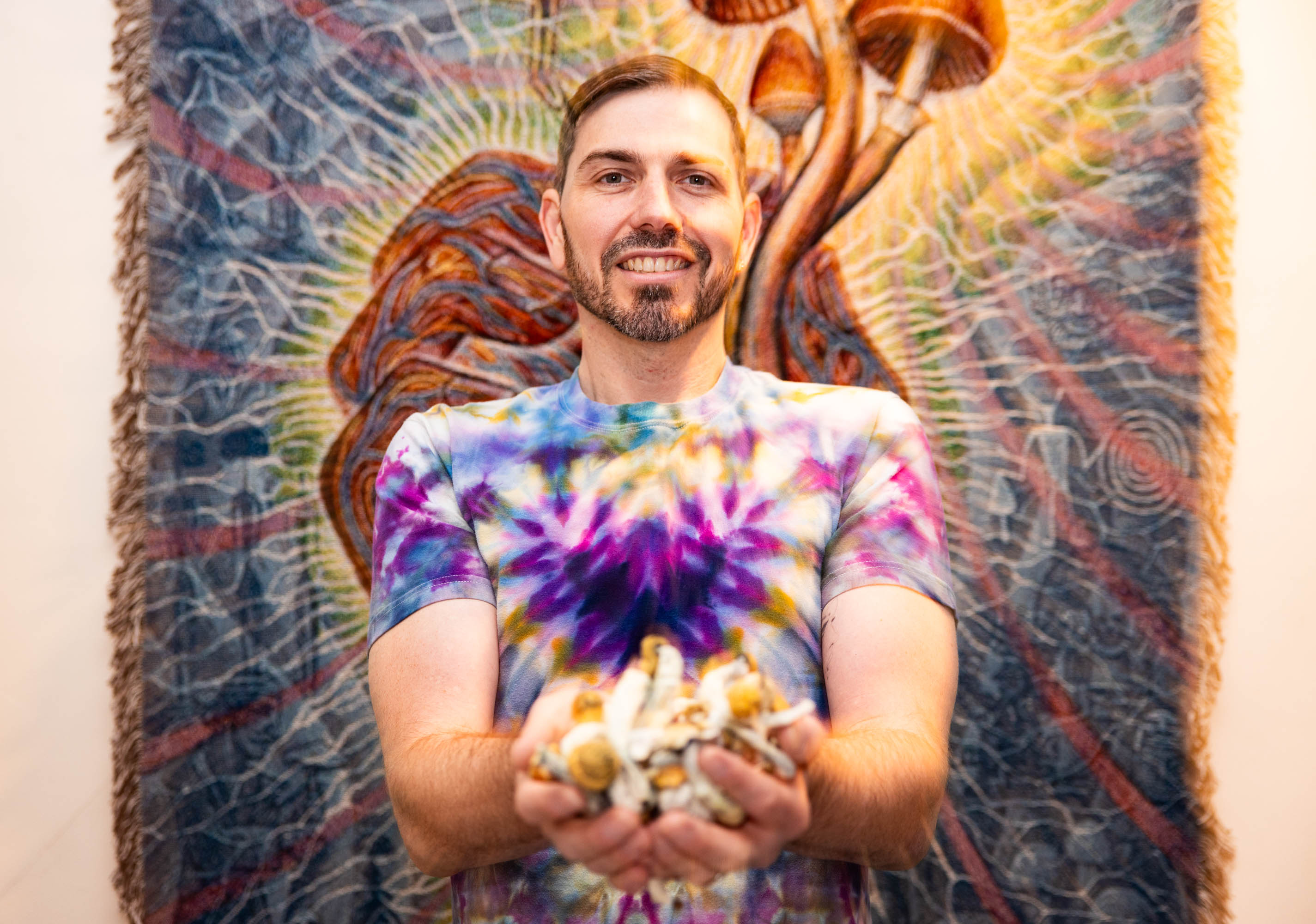 A smiling man in a tie-dye shirt holds magic mushrooms, standing before a colorful tapestry.